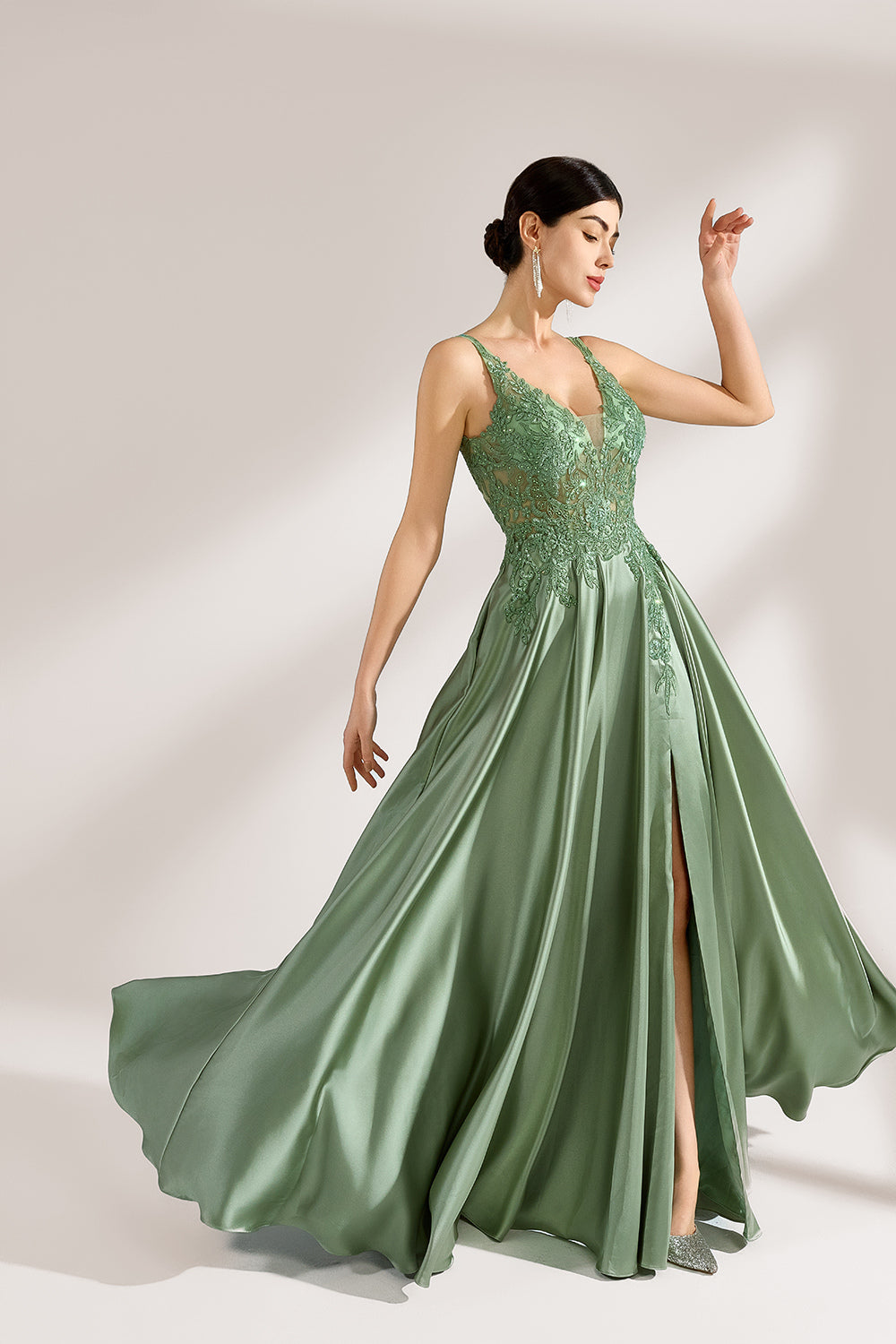 A woman with an elegant updo and a poised stance wears a sage - green satin gown. It has thin straps, a lace - detailed, V - neck bodice, and a high side slit. Paired with silver - tone heels, the outfit radiates grace, perfect for formal events like galas or weddings.1