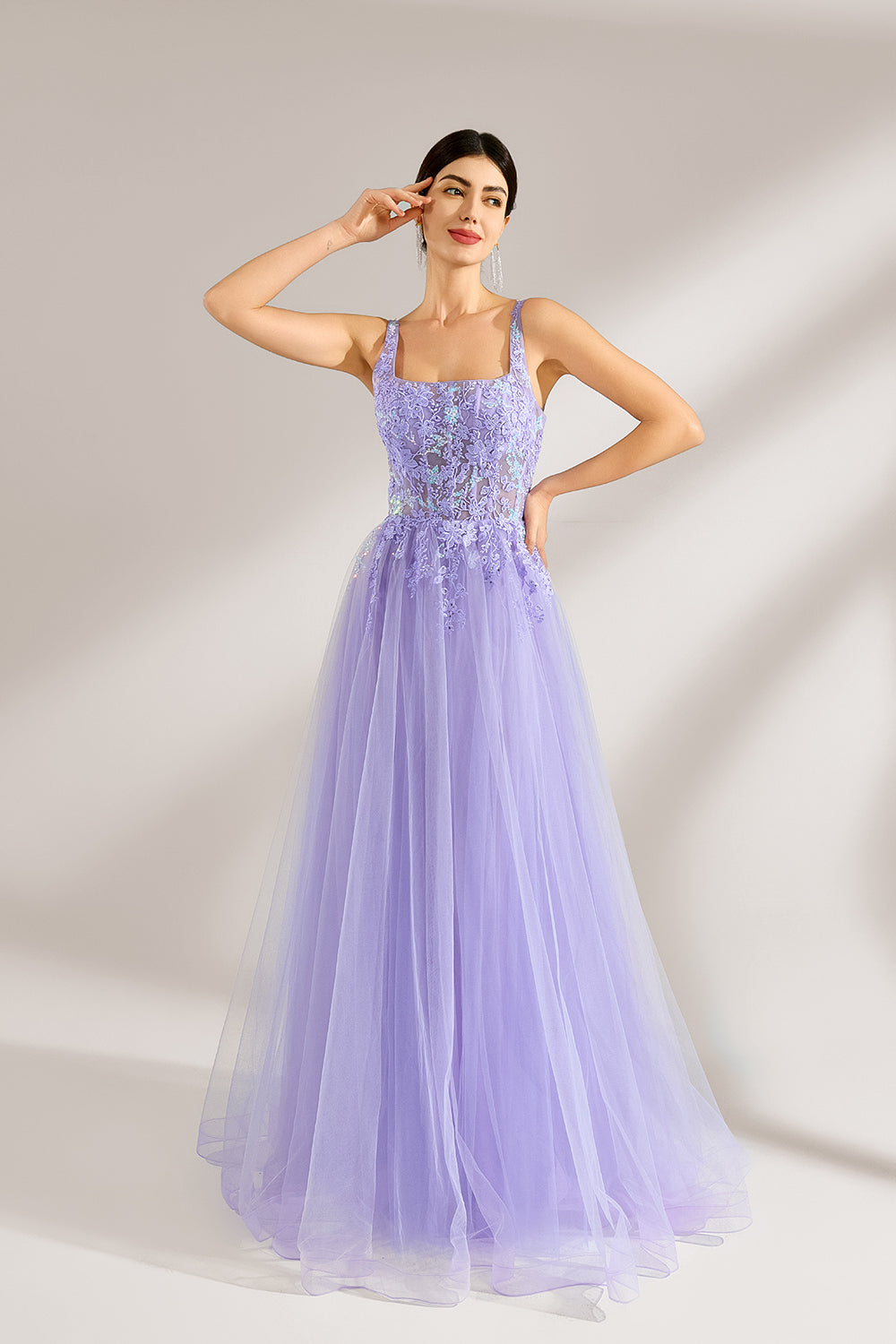The model has her hair neatly styled and wears long, elegant earrings. The dress is lavender. It features a square neckline with spaghetti straps, and the bodice is adorned with delicate lace. The skirt is made of flowing tulle, creating a dreamy and elegant look, perfect for formal occasions.1