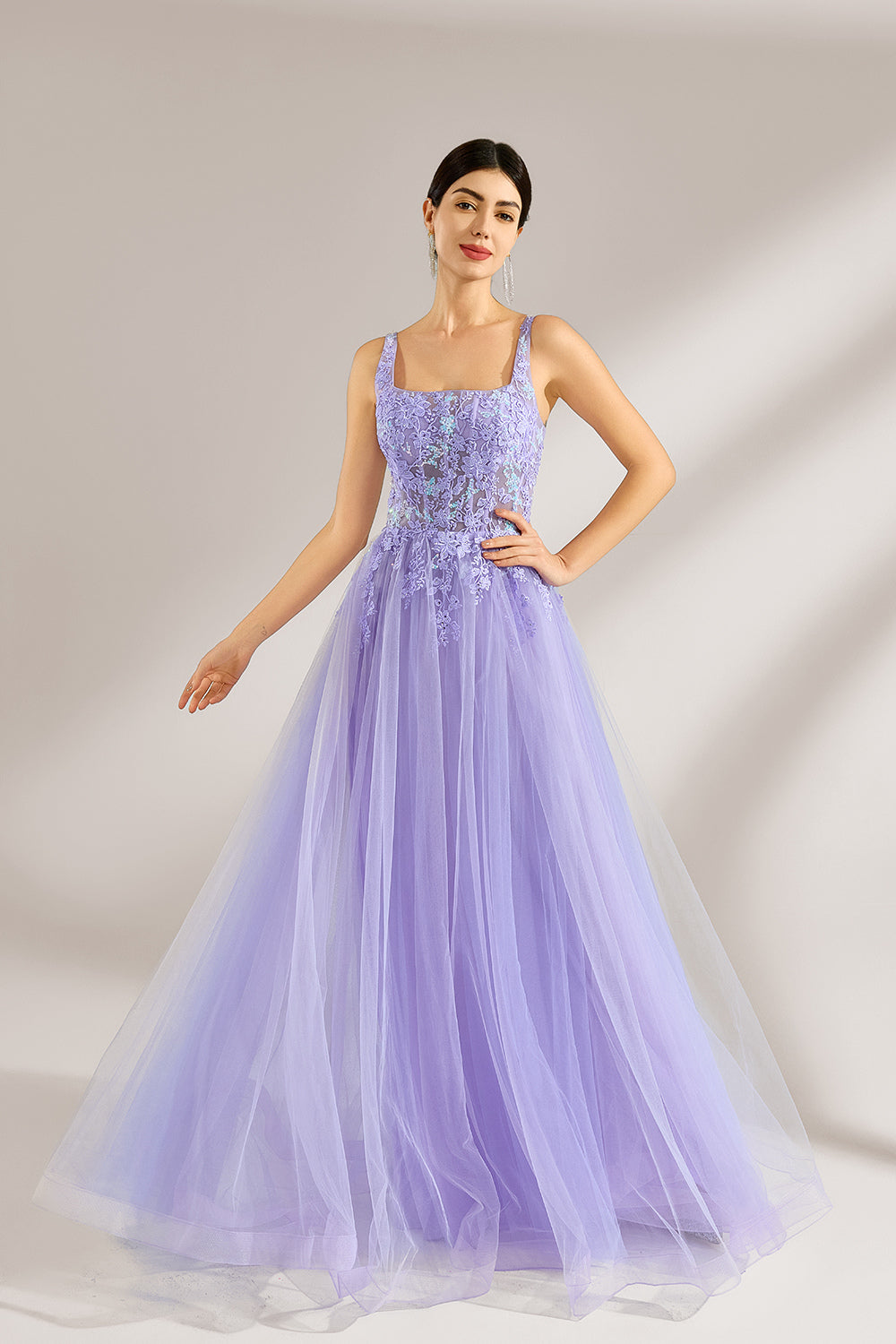 The model has her hair neatly styled and wears long, elegant earrings. The dress is lavender. It features a square neckline with spaghetti straps, and the bodice is adorned with delicate lace. The skirt is made of flowing tulle, creating a dreamy and elegant look, perfect for formal occasions.3