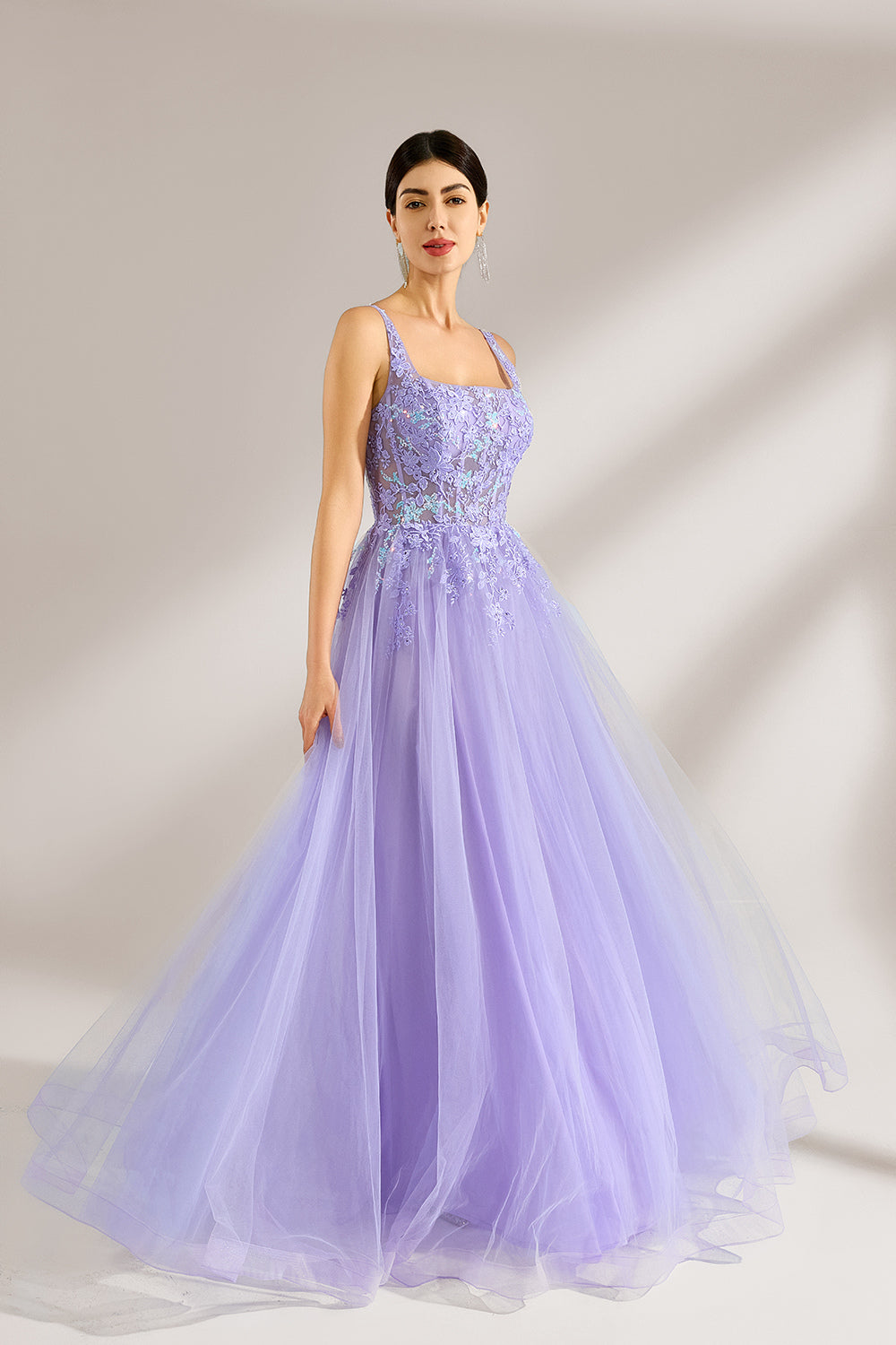 The model has her hair neatly styled and wears long, elegant earrings. The dress is lavender. It features a square neckline with spaghetti straps, and the bodice is adorned with delicate lace. The skirt is made of flowing tulle, creating a dreamy and elegant look, perfect for formal occasions.4