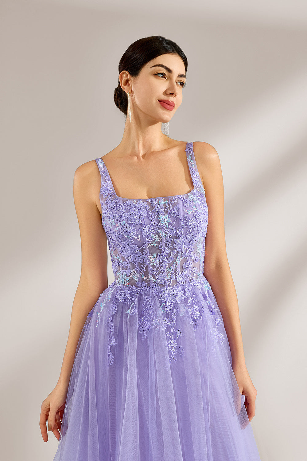 The model has her hair neatly styled and wears long, elegant earrings. The dress is lavender. It features a square neckline with spaghetti straps, and the bodice is adorned with delicate lace. The skirt is made of flowing tulle, creating a dreamy and elegant look, perfect for formal occasions.5