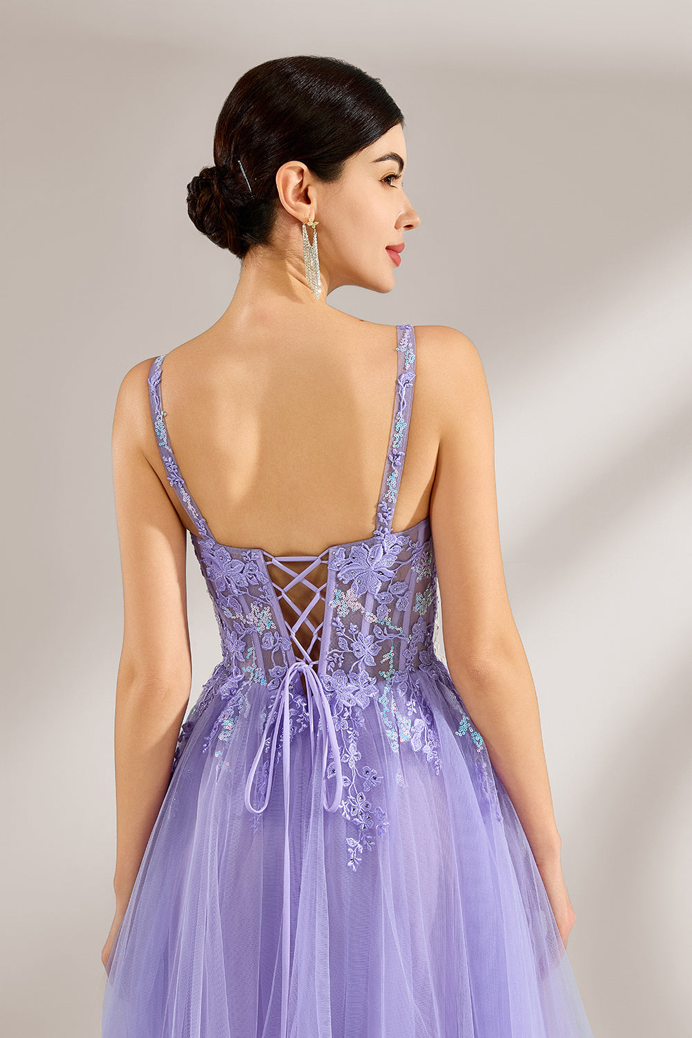The model has her hair neatly styled and wears long, elegant earrings. The dress is lavender. It features a square neckline with spaghetti straps, and the bodice is adorned with delicate lace. The skirt is made of flowing tulle, creating a dreamy and elegant look, perfect for formal occasions.6