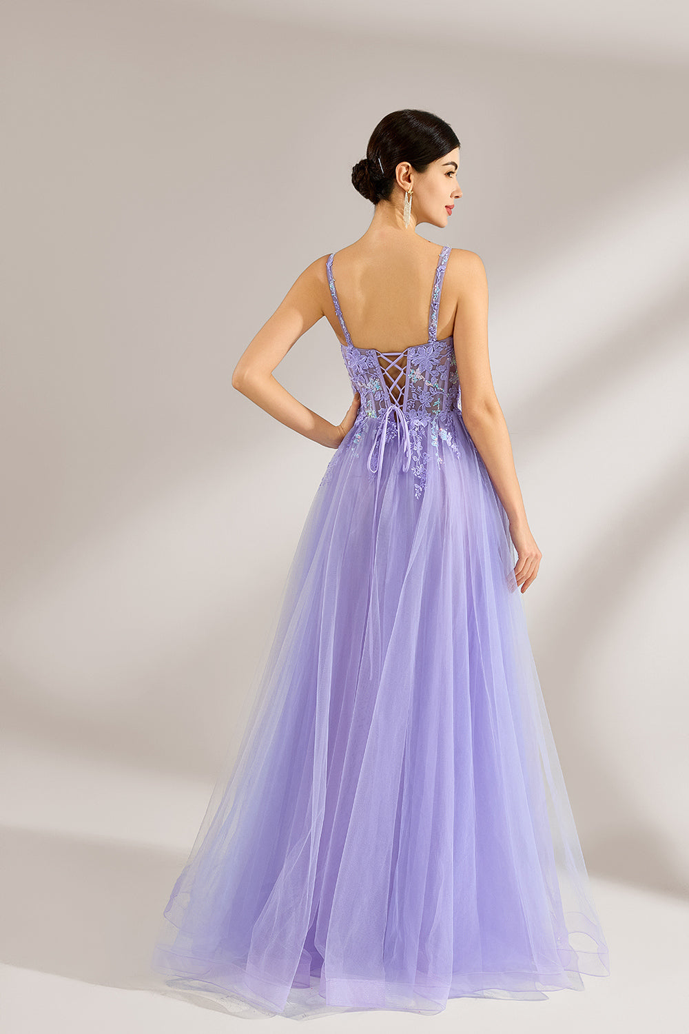 The model has her hair neatly styled and wears long, elegant earrings. The dress is lavender. It features a square neckline with spaghetti straps, and the bodice is adorned with delicate lace. The skirt is made of flowing tulle, creating a dreamy and elegant look, perfect for formal occasions.2