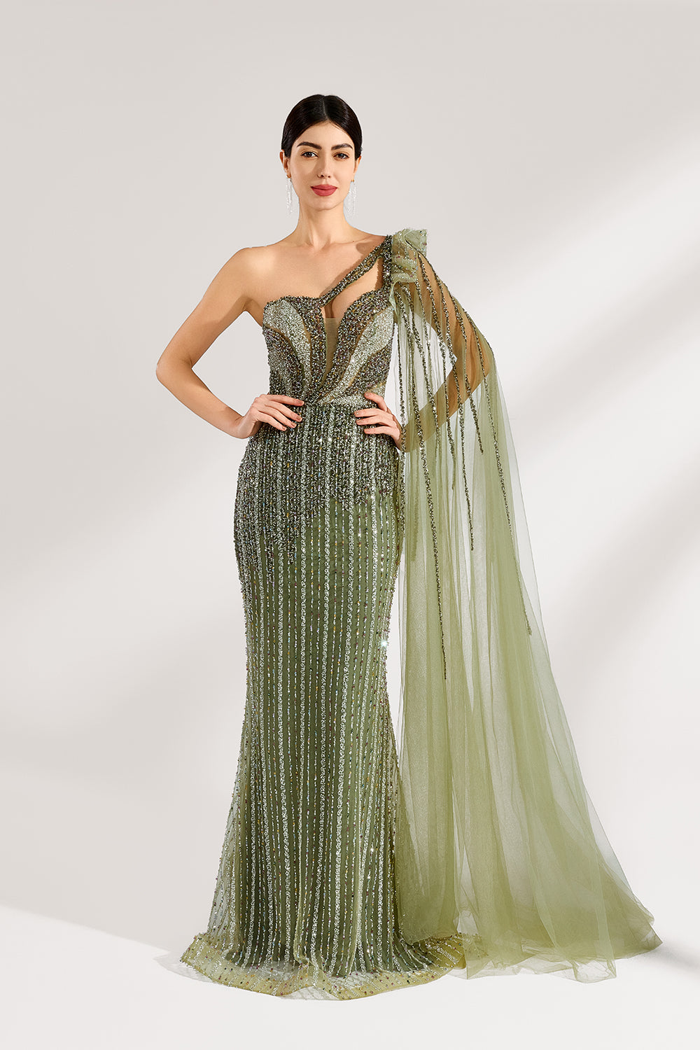Illuminated softly, a woman captivates in an olive - green one - shoulder evening gown. The sequined mermaid silhouette, paired with a tulle cape, creates a striking and elegant look for formal events.1