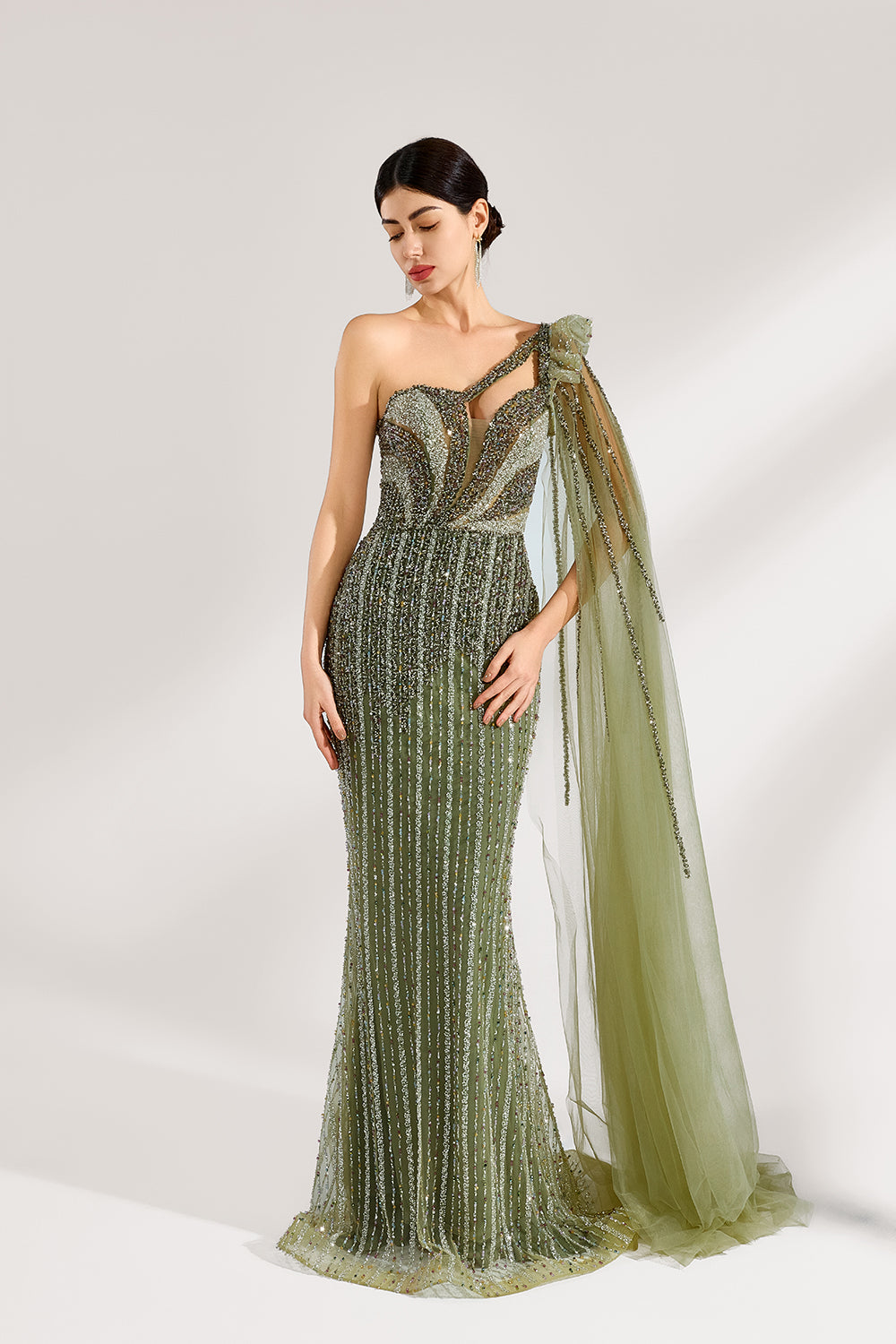 Illuminated softly, a woman captivates in an olive - green one - shoulder evening gown. The sequined mermaid silhouette, paired with a tulle cape, creates a striking and elegant look for formal events.4