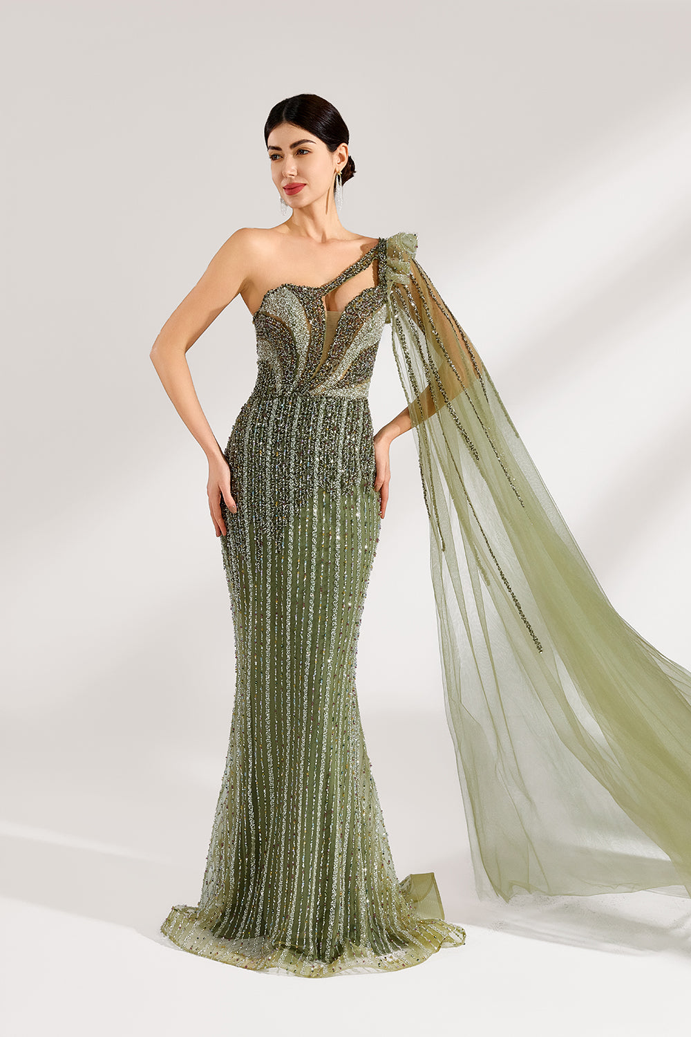 Illuminated softly, a woman captivates in an olive - green one - shoulder evening gown. The sequined mermaid silhouette, paired with a tulle cape, creates a striking and elegant look for formal events.5