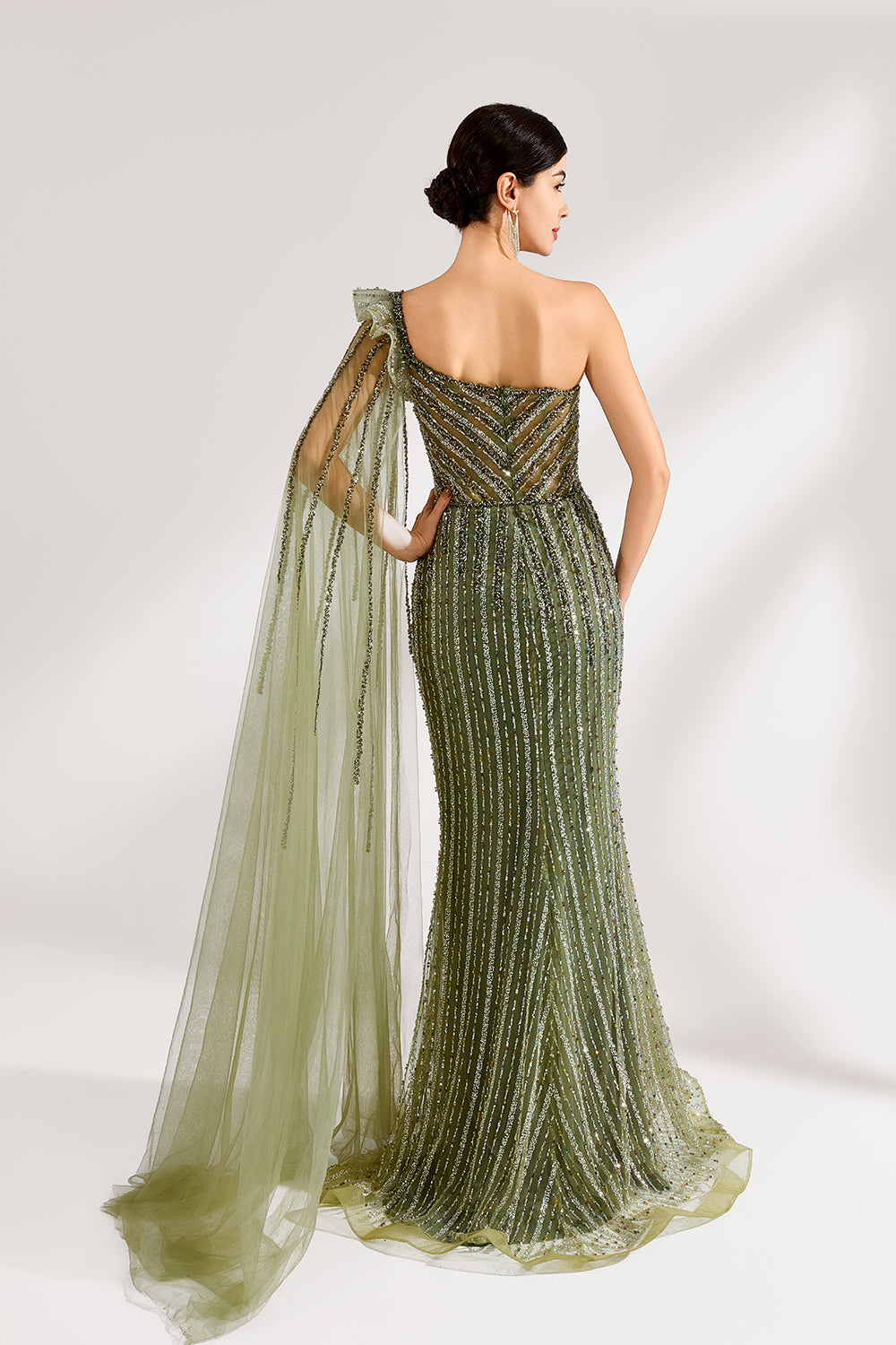 Illuminated softly, a woman captivates in an olive - green one - shoulder evening gown. The sequined mermaid silhouette, paired with a tulle cape, creates a striking and elegant look for formal events.2