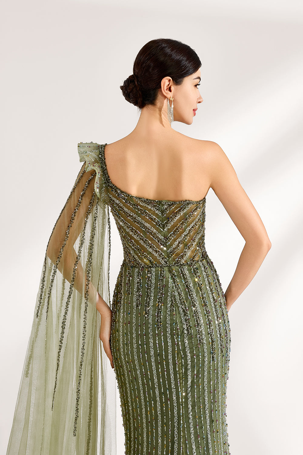 Illuminated softly, a woman captivates in an olive - green one - shoulder evening gown. The sequined mermaid silhouette, paired with a tulle cape, creates a striking and elegant look for formal events.6