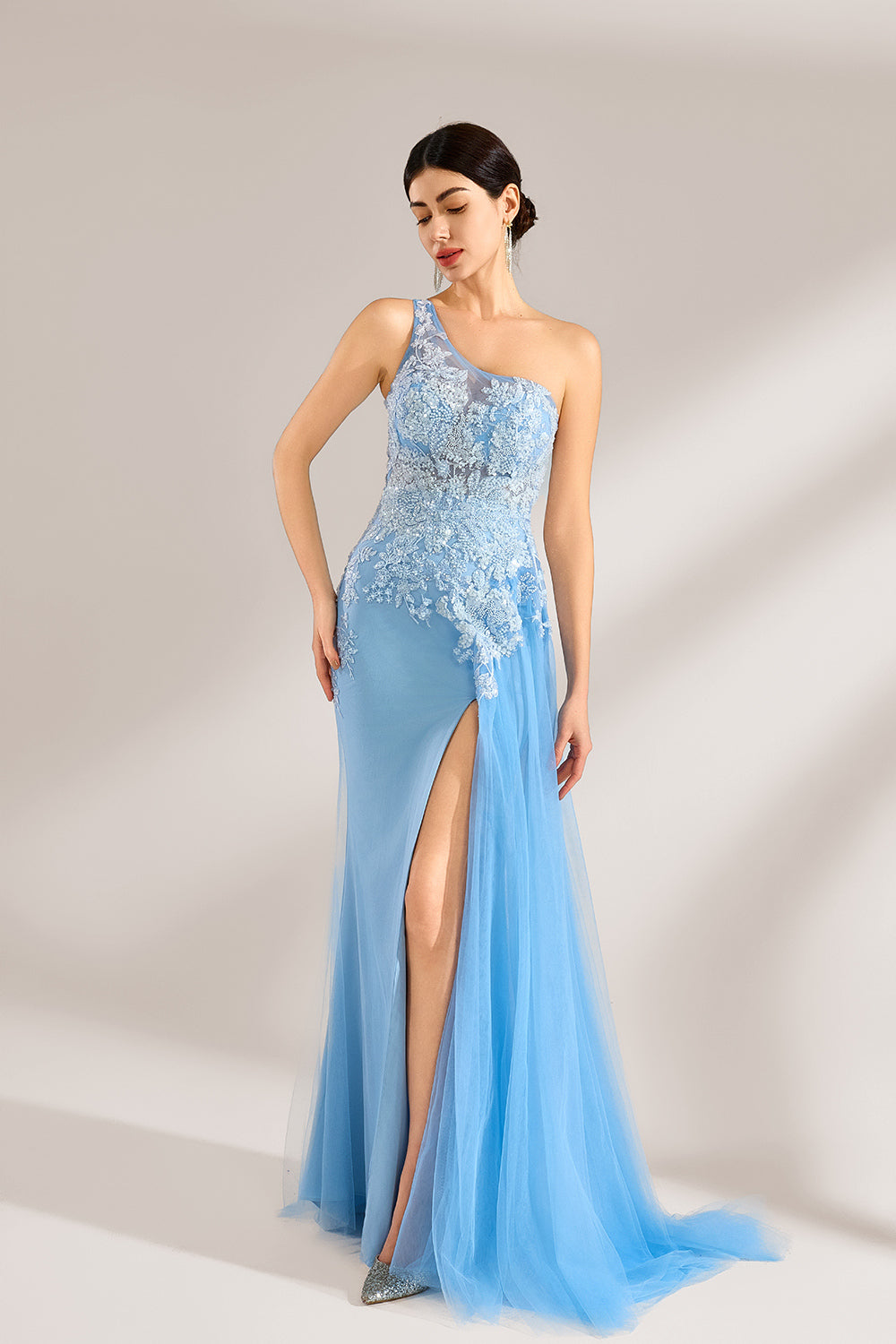 A woman with sleek dark hair and a poised stance wears a light - blue tulle gown. It has a one - shoulder design, lace - detailed bodice, and a high side slit. Paired with silver heels, the outfit radiates elegance, perfect for formal events like galas, blending romance and glamour.4