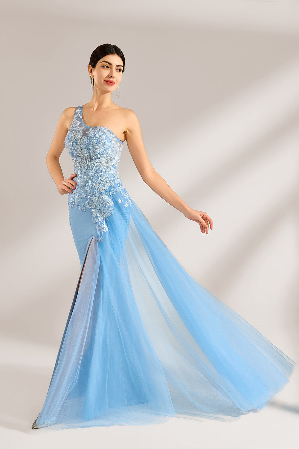 A woman with sleek dark hair and a poised stance wears a light - blue tulle gown. It has a one - shoulder design, lace - detailed bodice, and a high side slit. Paired with silver heels, the outfit radiates elegance, perfect for formal events like galas, blending romance and glamour.1