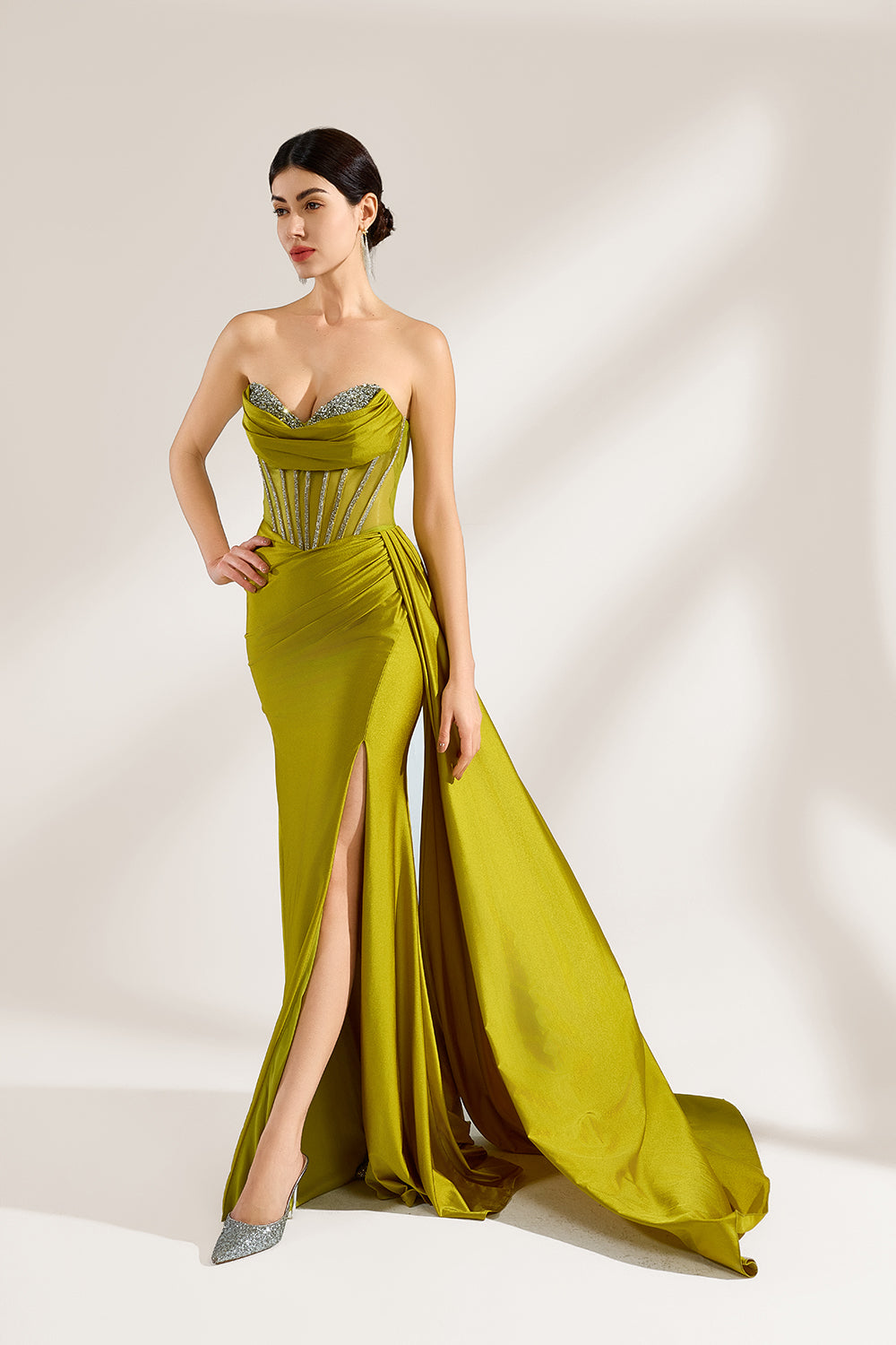 A woman stands against a light background, wearing an olive - green satin gown. It features a strapless neckline with crystal embellishments, a figure - hugging silhouette, a high side slit, and a dramatic train. The elegant details create a glamorous look, perfect for formal events like galas or red - carpet occasions.