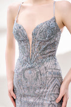 A woman stands against a light - hued background, wearing a light - grey, sheer gown adorned with intricate silver - toned bead patterns. It has thin straps, a deep V - neckline, and a mermaid silhouette. The detailed beadwork and sheer fabric create a glamorous look, perfect for proms or formal parties, radiating sophistication.6