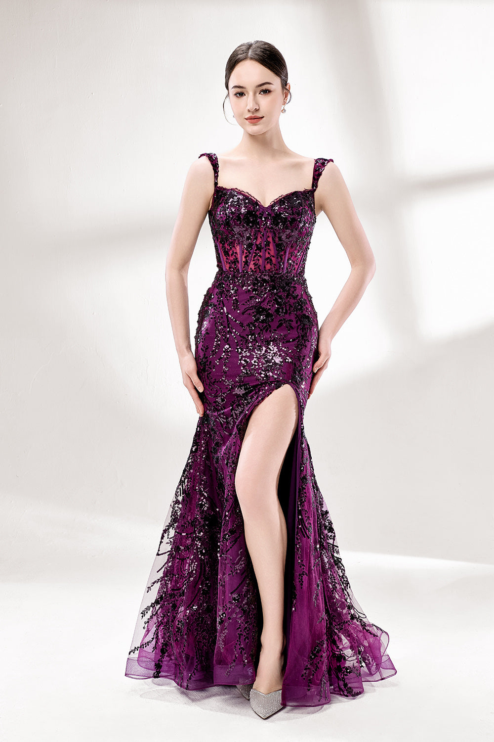Set against a light background, a woman wears a purple tulle gown embellished with sequin embroidery. It has thin straps, a sweetheart neckline, a sheer bodice patterned with florals, and a mermaid skirt with a high side slit. Silver heels complete the look, with the sparkling sequins and refined silhouette ideal for proms or formal dinners.1