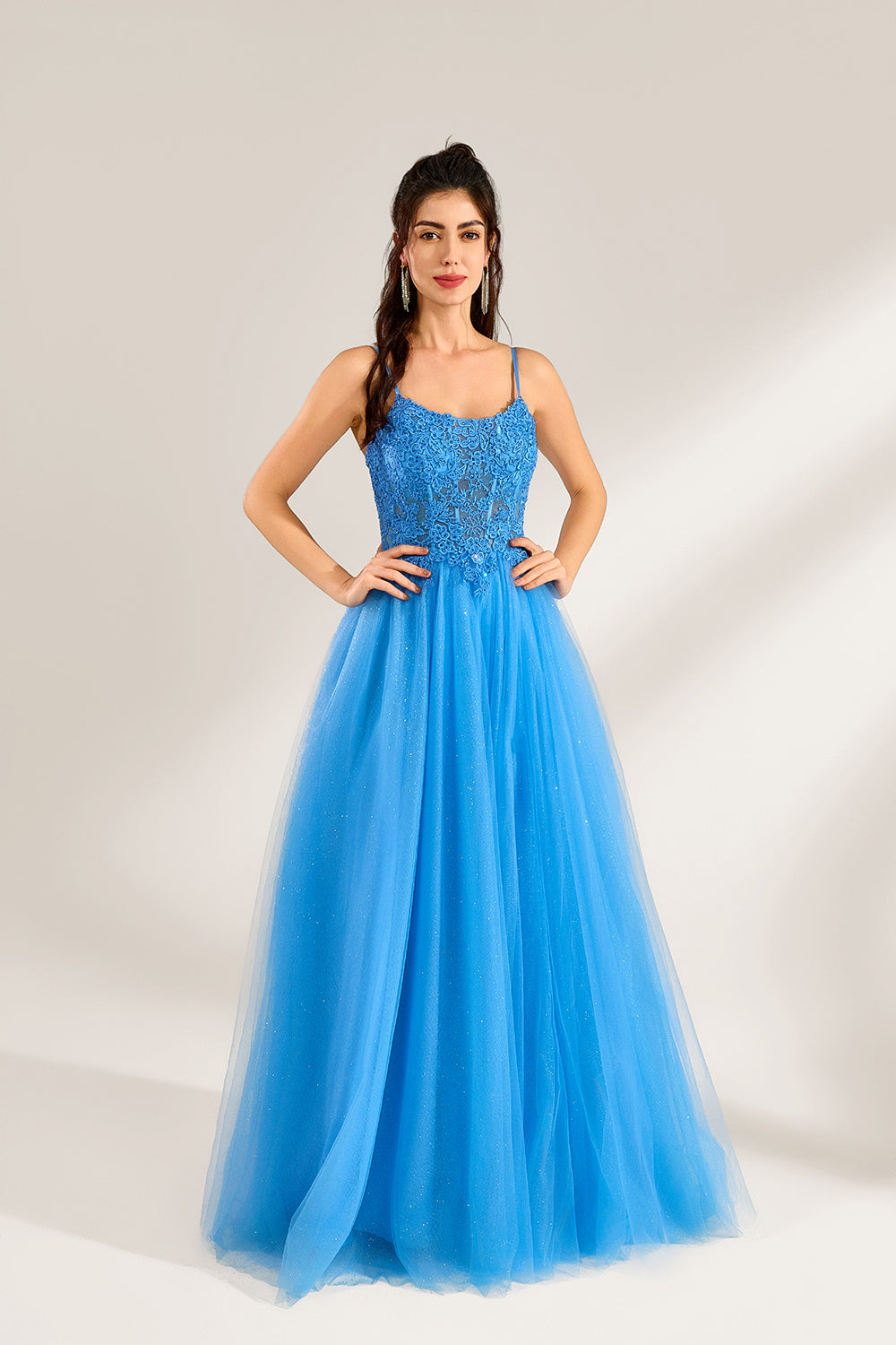 A woman stands against a light background, wearing a blue tulle gown. It features thin straps, a square neckline, intricate lace embroidery on the bodice, and a voluminous tulle skirt with subtle glitter. The elegant design creates a romantic and glamorous look, perfect for formal events like proms or weddings.4
