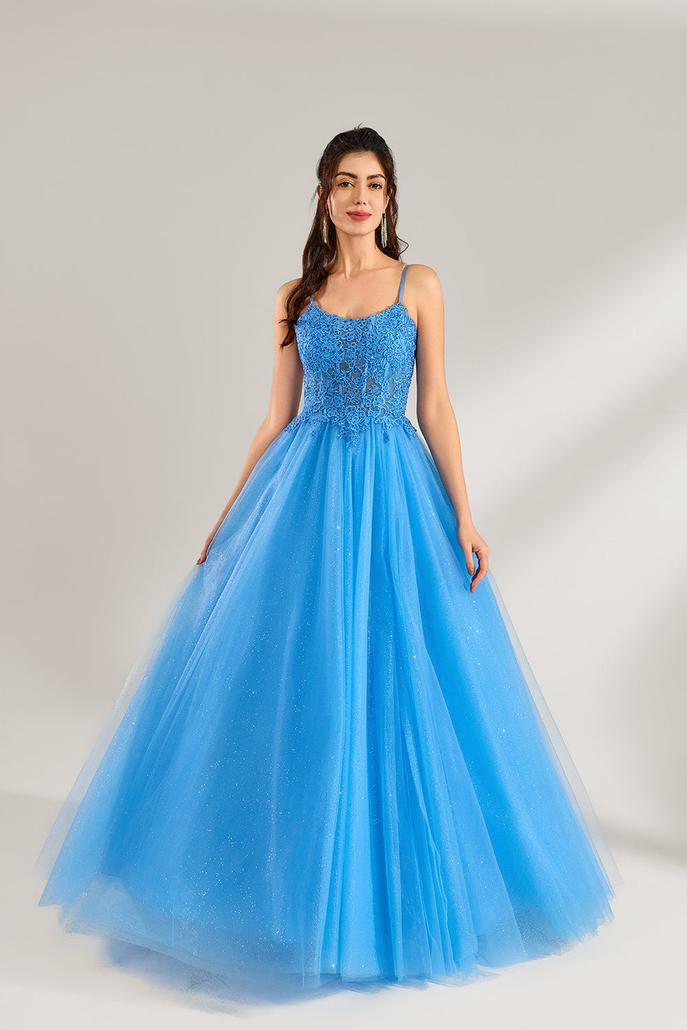 A woman stands against a light background, wearing a blue tulle gown. It features thin straps, a square neckline, intricate lace embroidery on the bodice, and a voluminous tulle skirt with subtle glitter. The elegant design creates a romantic and glamorous look, perfect for formal events like proms or weddings.1