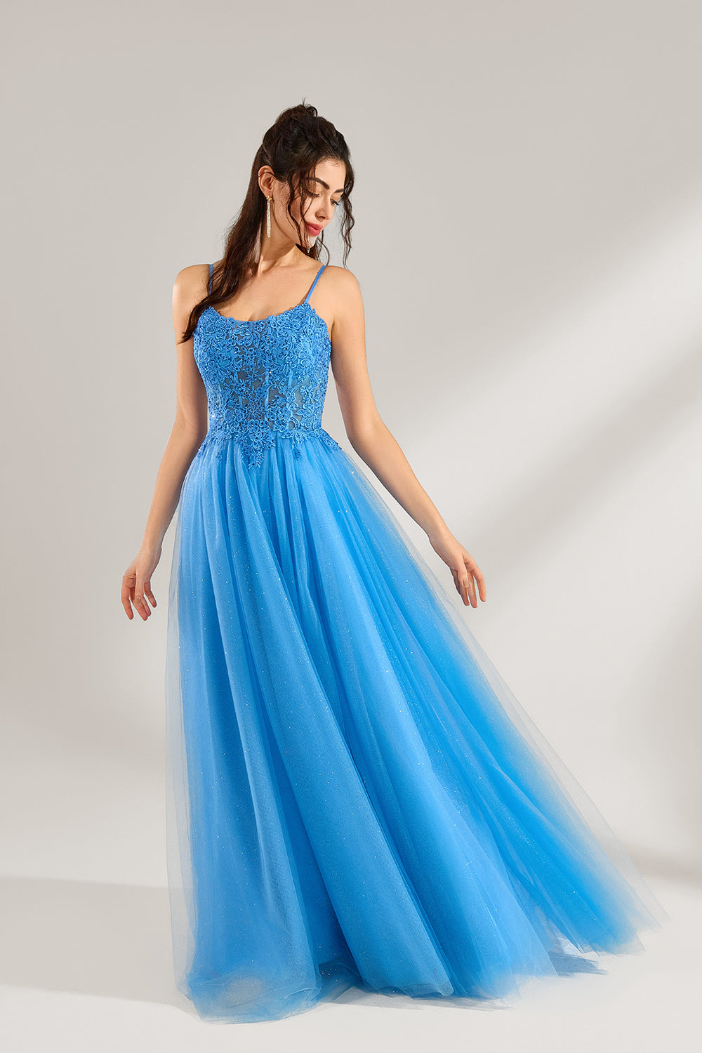 A woman stands against a light background, wearing a blue tulle gown. It features thin straps, a square neckline, intricate lace embroidery on the bodice, and a voluminous tulle skirt with subtle glitter. The elegant design creates a romantic and glamorous look, perfect for formal events like proms or weddings.3