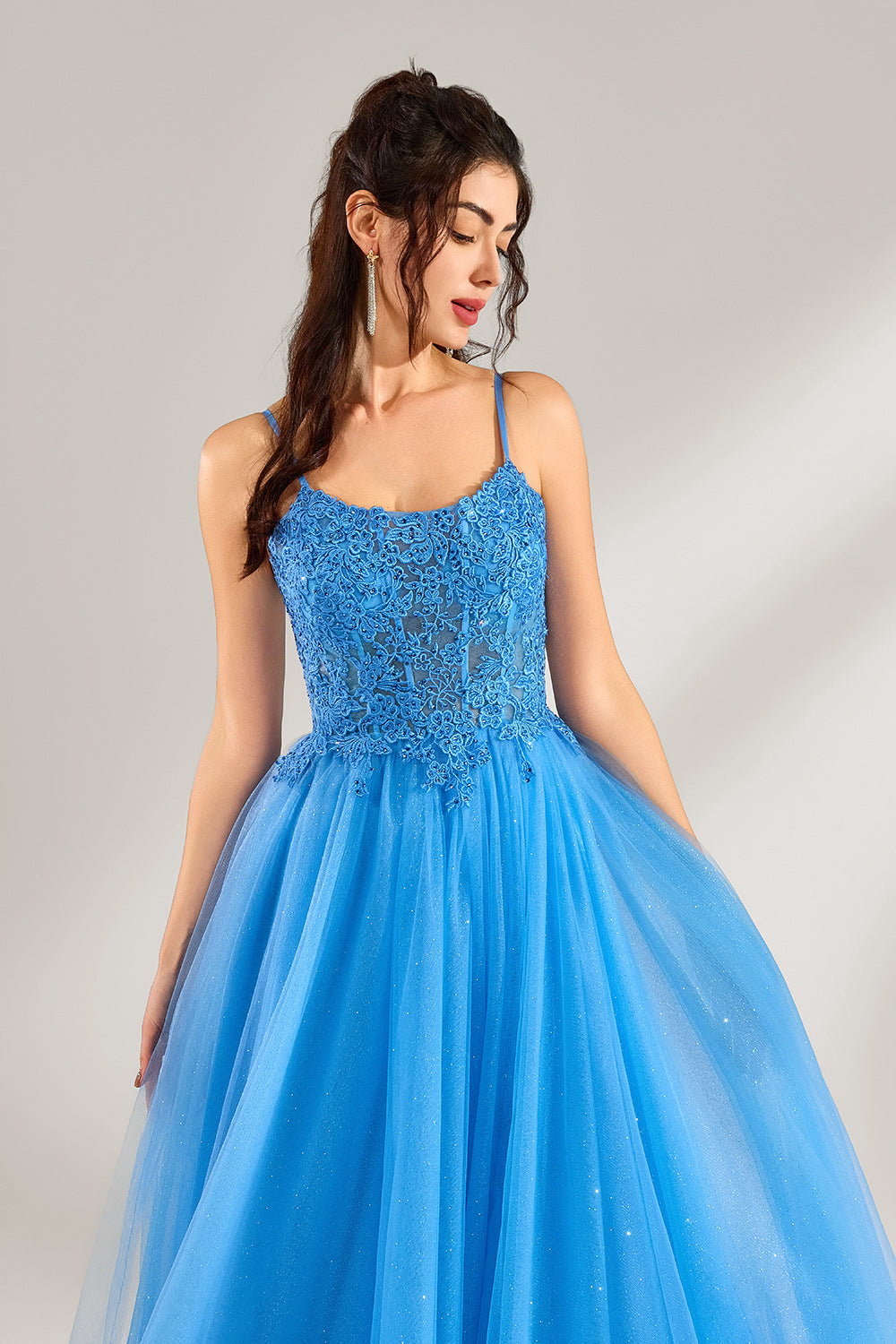 A woman stands against a light background, wearing a blue tulle gown. It features thin straps, a square neckline, intricate lace embroidery on the bodice, and a voluminous tulle skirt with subtle glitter. The elegant design creates a romantic and glamorous look, perfect for formal events like proms or weddings.5