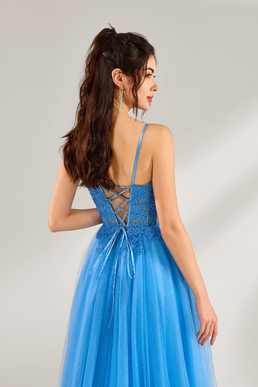 A woman stands against a light background, wearing a blue tulle gown. It features thin straps, a square neckline, intricate lace embroidery on the bodice, and a voluminous tulle skirt with subtle glitter. The elegant design creates a romantic and glamorous look, perfect for formal events like proms or weddings.6