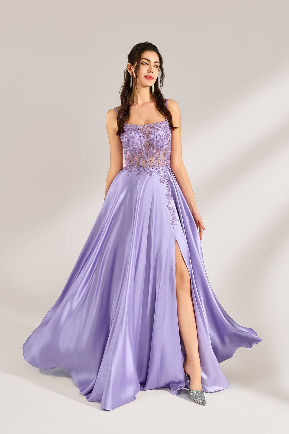 A woman with long, wavy dark hair and a confident pose wears a light - purple satin gown. It has thin straps, a sheer lace - detailed bodice, and a high side slit. Paired with silver - tone heels, the outfit radiates elegance, perfect for formal events like galas or evening receptions.1