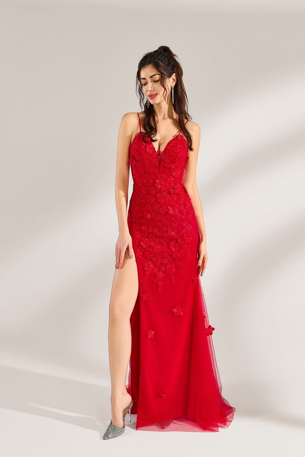 The model has her long hair tied up in a neat style and wears elegant earrings. The dress is a striking red color. It features spaghetti straps, a deep V-neck, and is adorned with delicate floral patterns. There's a high side slit that adds a touch of allure, and the tulle skirt flows gracefully. It's perfect for formal occasions like galas or weddings.4