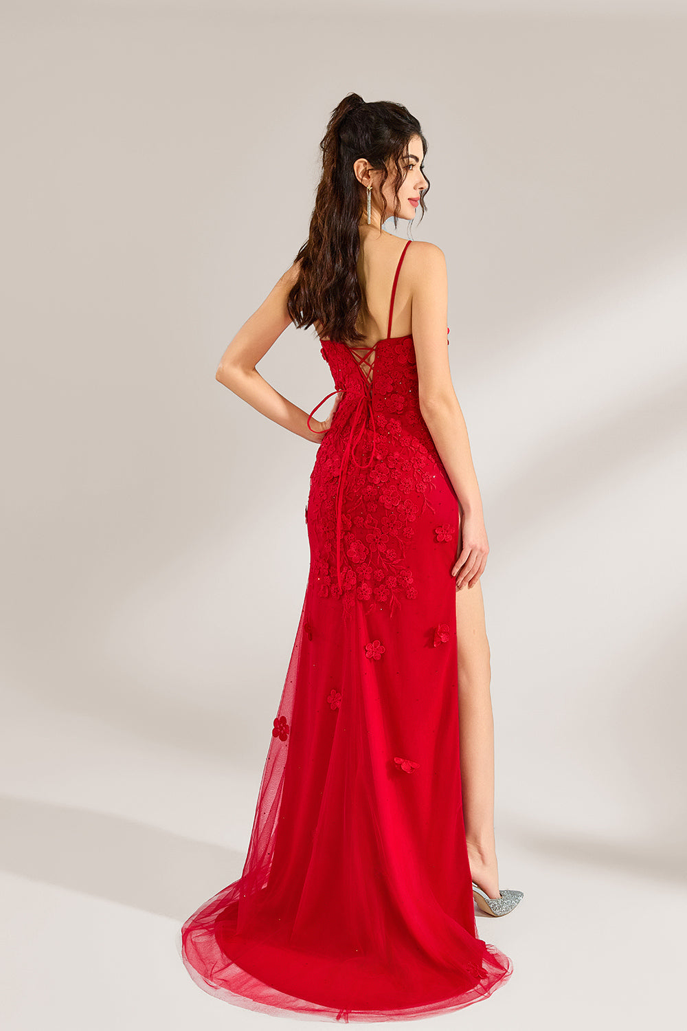 The model has her long hair tied up in a neat style and wears elegant earrings. The dress is a striking red color. It features spaghetti straps, a deep V-neck, and is adorned with delicate floral patterns. There's a high side slit that adds a touch of allure, and the tulle skirt flows gracefully. It's perfect for formal occasions like galas or weddings.2