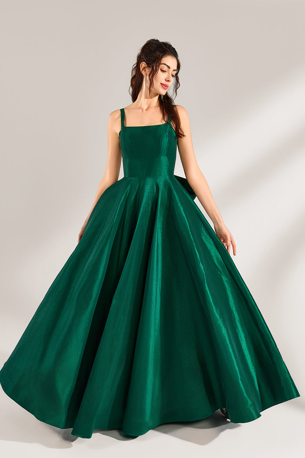 A woman stands against a light background, wearing a dark - green satin gown. It features square neckline, wide straps, and a bow at the waist. The voluminous skirt and elegant silhouette exude sophistication, perfect for formal events like galas or weddings, radiating timeless charm.1