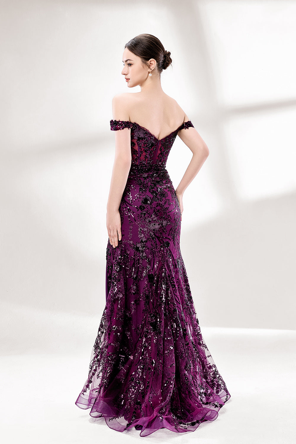 Set against a light background, a woman wears a purple tulle gown embellished with sequin embroidery. It has thin straps, a sweetheart neckline, a sheer bodice patterned with florals, and a mermaid skirt with a high side slit. Silver heels complete the look, with the sparkling sequins and refined silhouette ideal for proms or formal dinners.2