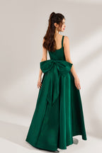 A woman stands against a light background, wearing a dark - green satin gown. It features square neckline, wide straps, and a bow at the waist. The voluminous skirt and elegant silhouette exude sophistication, perfect for formal events like galas or weddings, radiating timeless charm.2