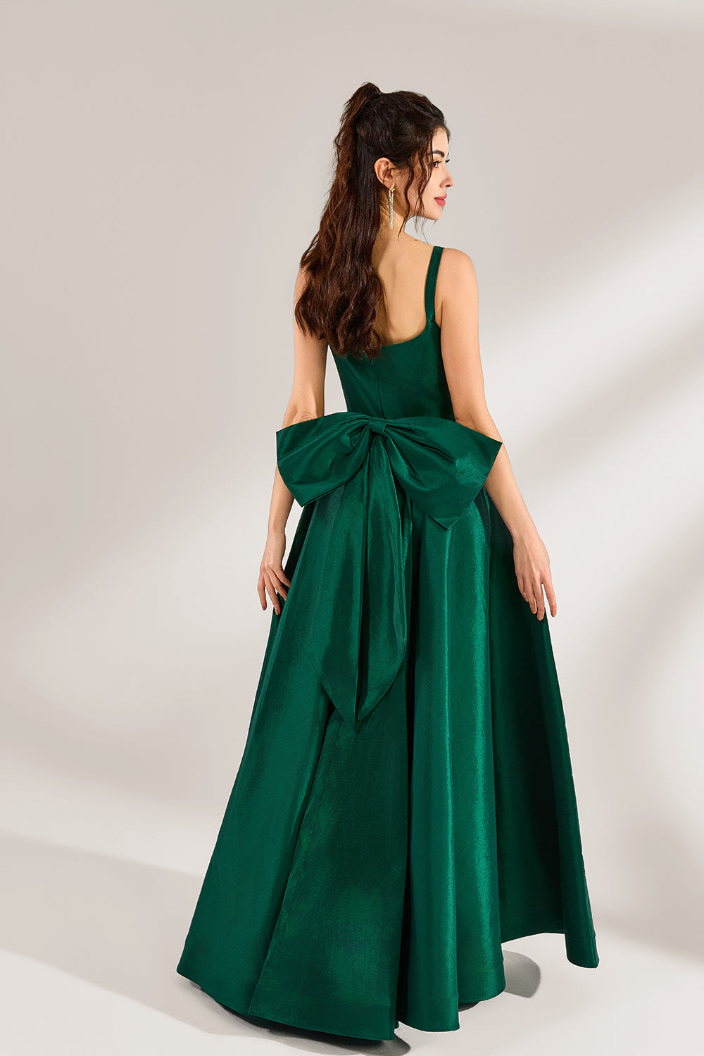 A woman stands against a light background, wearing a dark - green satin gown. It features square neckline, wide straps, and a bow at the waist. The voluminous skirt and elegant silhouette exude sophistication, perfect for formal events like galas or weddings, radiating timeless charm.2