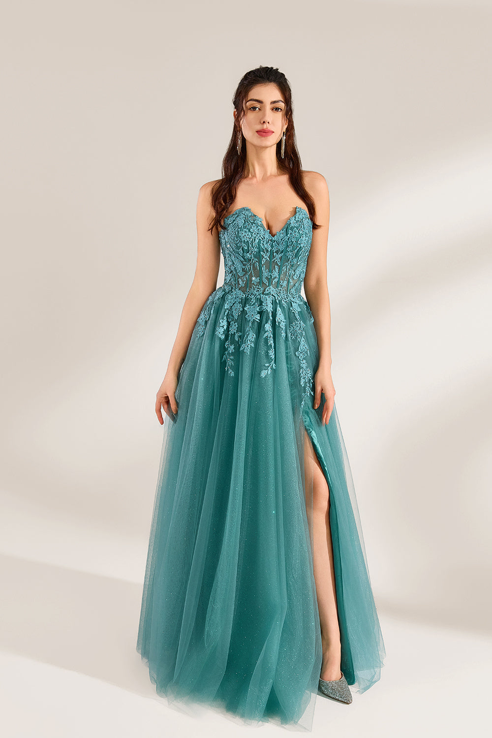 In front of a light - colored background, a woman is dressed in a teal tulle strapless gown. It has a sweetheart neckline, with the bodice adorned with delicate lace embroidery, and a high side slit. The voluminous tulle skirt, which has a slight shimmer, gives an elegant and romantic appearance, ideal for formal events such as proms or weddings.1