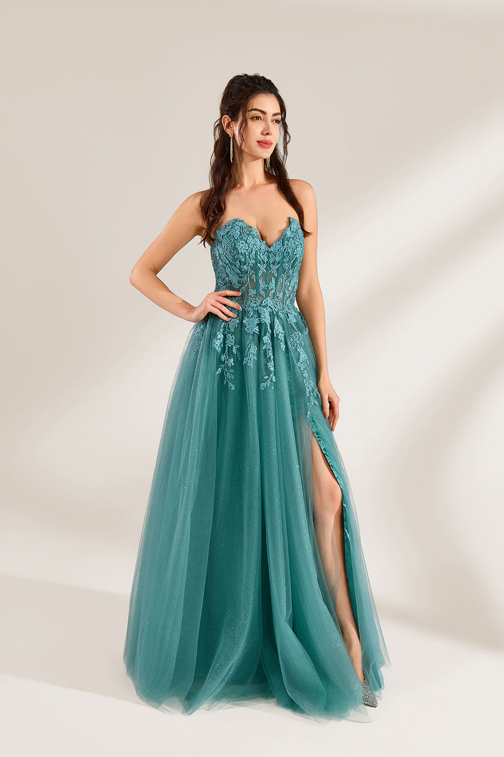 In front of a light - colored background, a woman is dressed in a teal tulle strapless gown. It has a sweetheart neckline, with the bodice adorned with delicate lace embroidery, and a high side slit. The voluminous tulle skirt, which has a slight shimmer, gives an elegant and romantic appearance, ideal for formal events such as proms or weddings.3