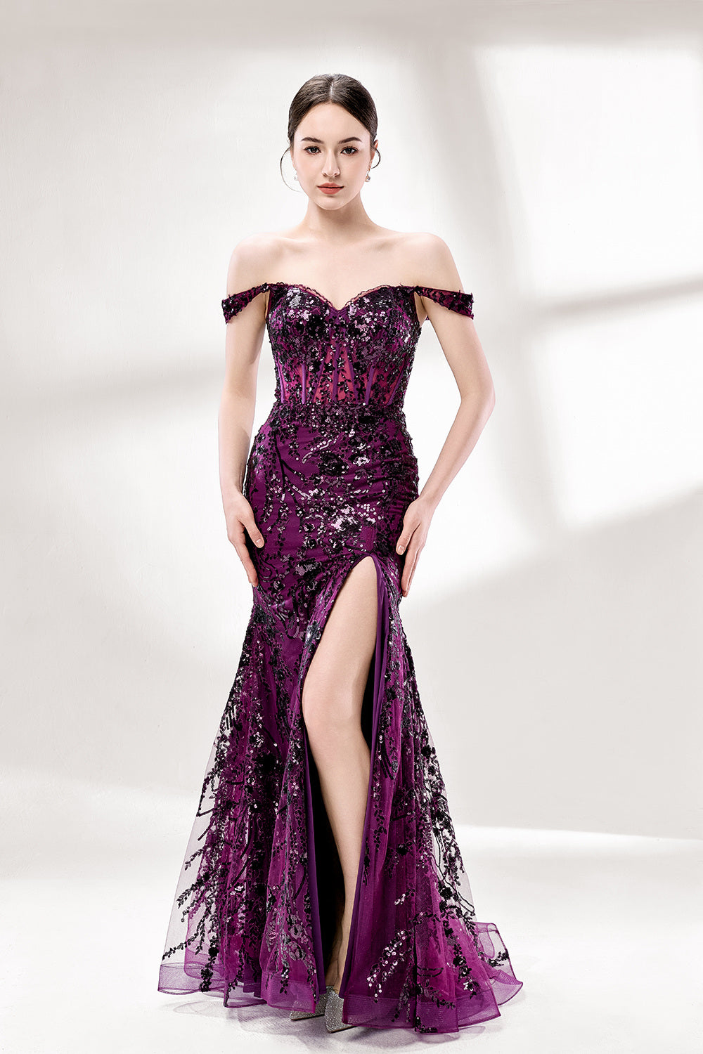 Set against a light background, a woman wears a purple tulle gown embellished with sequin embroidery. It has thin straps, a sweetheart neckline, a sheer bodice patterned with florals, and a mermaid skirt with a high side slit. Silver heels complete the look, with the sparkling sequins and refined silhouette ideal for proms or formal dinners.5