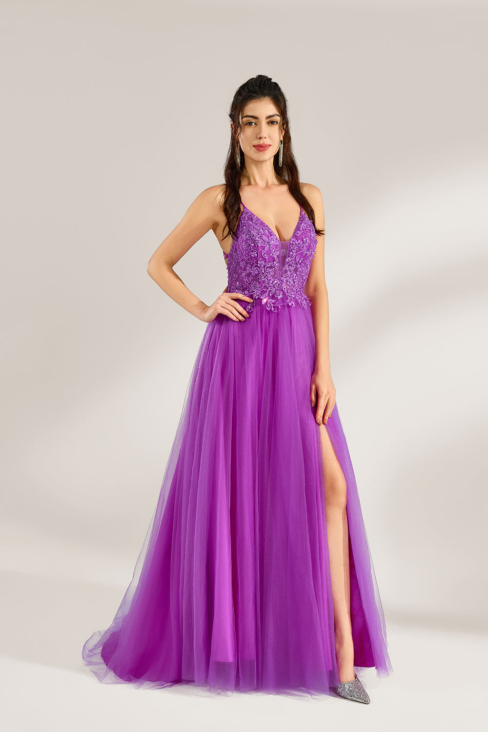 Set against a light background, a woman adorns a purple tulle gown. It has thin straps, a deep V - neckline with delicate lace embroidery on the bodice, and a high side slit. The flowing tulle skirt gives off an elegant and romantic vibe, ideal for formal events such as proms or weddings.1