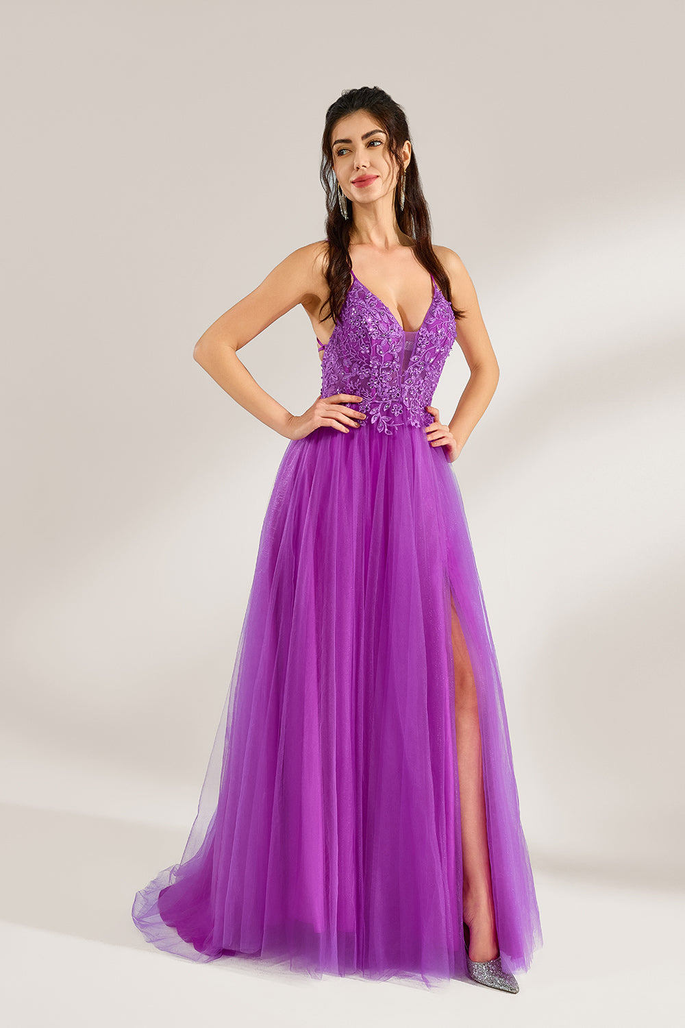 Set against a light background, a woman adorns a purple tulle gown. It has thin straps, a deep V - neckline with delicate lace embroidery on the bodice, and a high side slit. The flowing tulle skirt gives off an elegant and romantic vibe, ideal for formal events such as proms or weddings.3