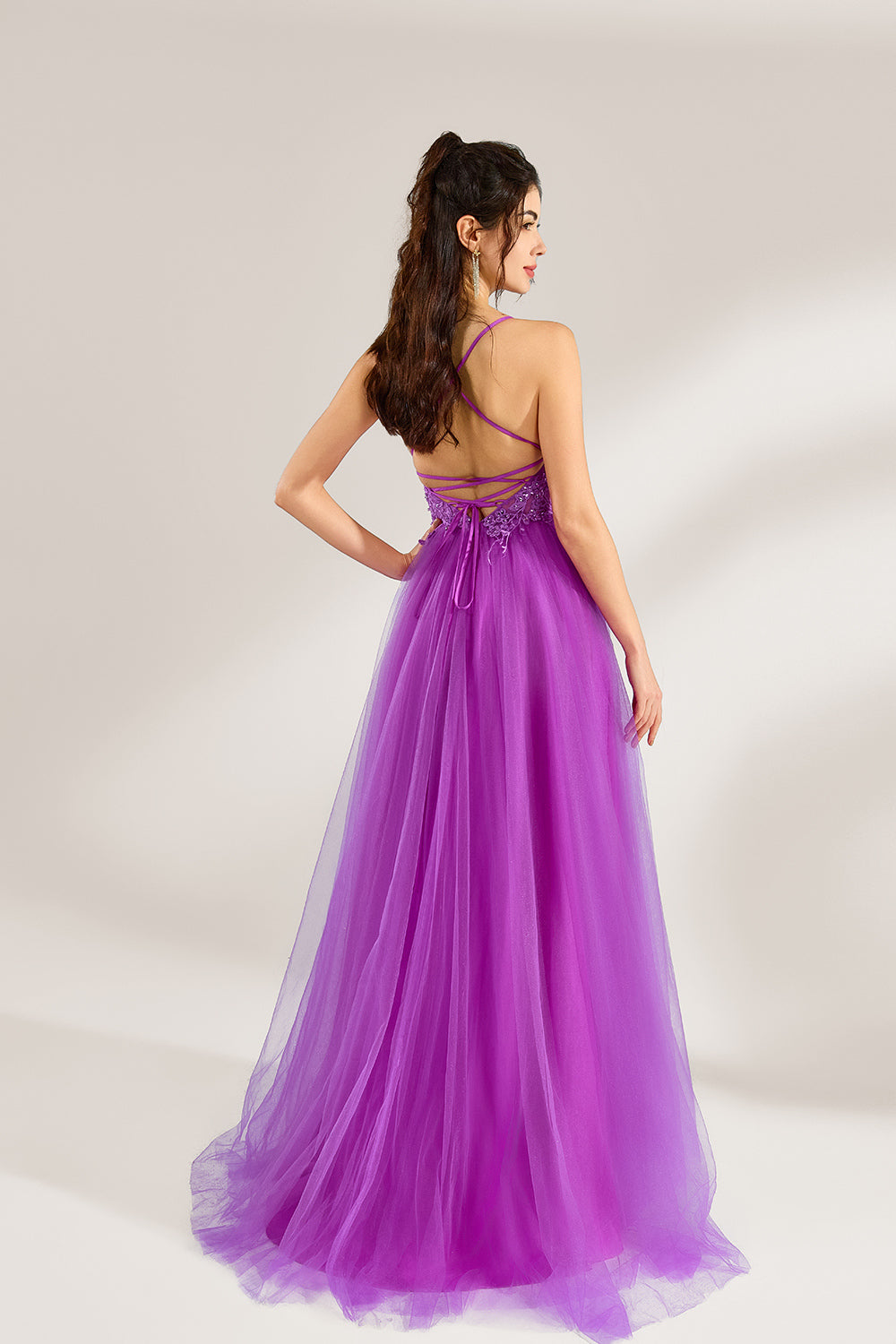 Set against a light background, a woman adorns a purple tulle gown. It has thin straps, a deep V - neckline with delicate lace embroidery on the bodice, and a high side slit. The flowing tulle skirt gives off an elegant and romantic vibe, ideal for formal events such as proms or weddings.2