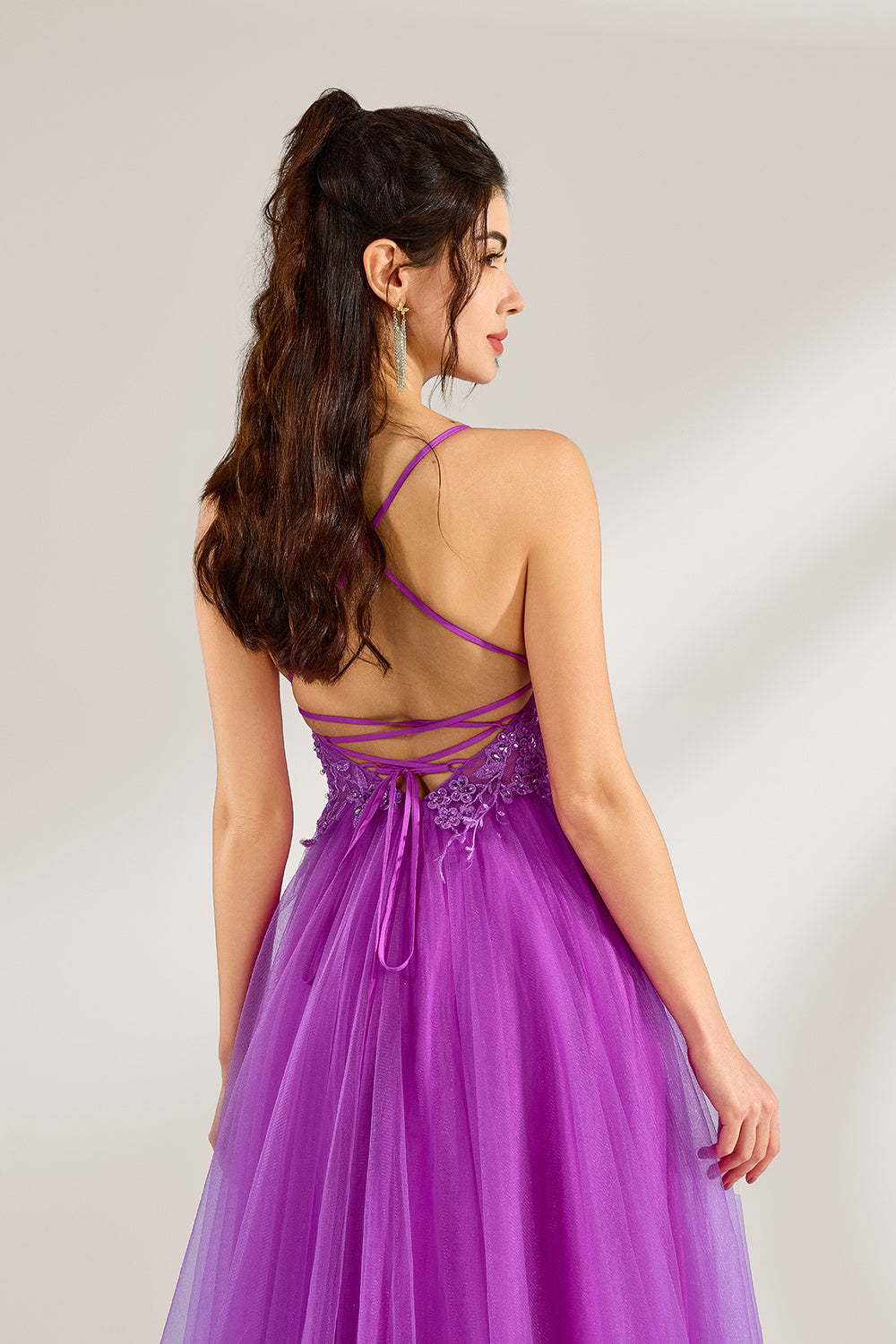 Set against a light background, a woman adorns a purple tulle gown. It has thin straps, a deep V - neckline with delicate lace embroidery on the bodice, and a high side slit. The flowing tulle skirt gives off an elegant and romantic vibe, ideal for formal events such as proms or weddings.5
