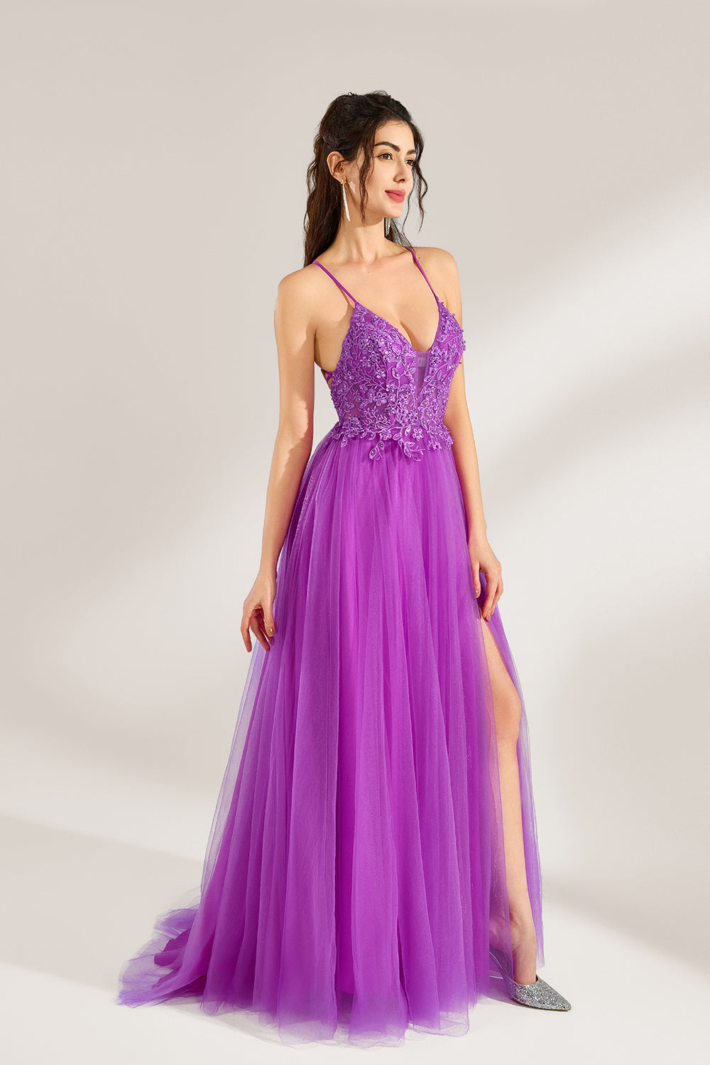 Set against a light background, a woman adorns a purple tulle gown. It has thin straps, a deep V - neckline with delicate lace embroidery on the bodice, and a high side slit. The flowing tulle skirt gives off an elegant and romantic vibe, ideal for formal events such as proms or weddings.6