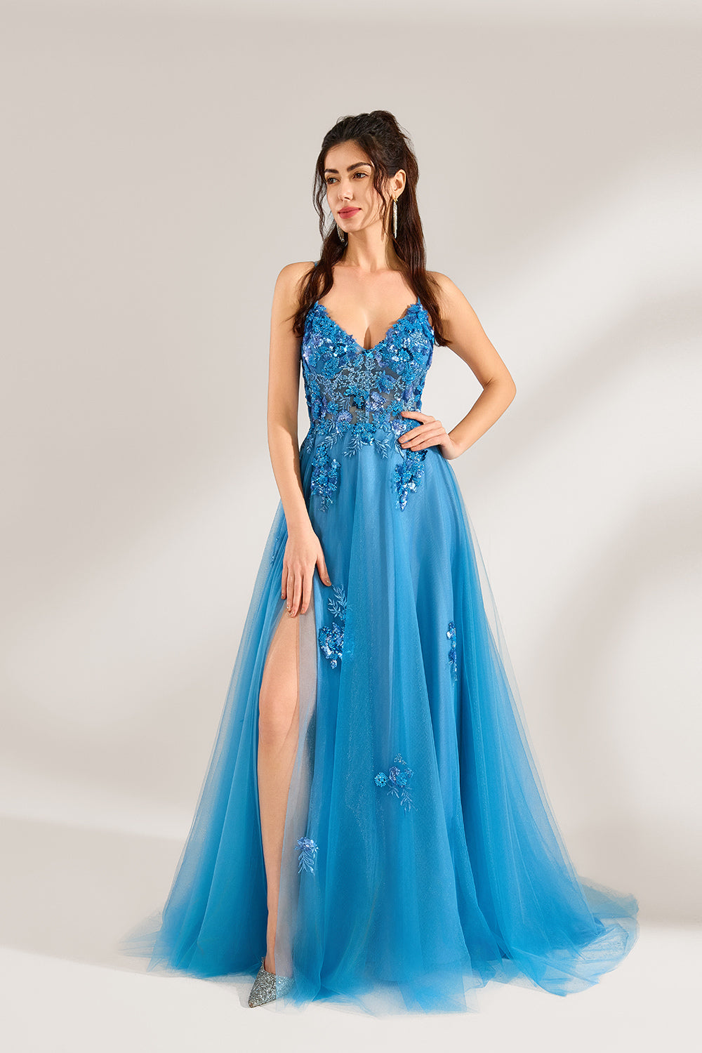 A woman stands against a light background, wearing a blue tulle gown. It features thin straps, a deep V - neckline, intricate floral appliqués with sequins on the bodice, and a high side slit. The voluminous tulle skirt creates a romantic and glamorous look, perfect for formal events like proms or weddings.1