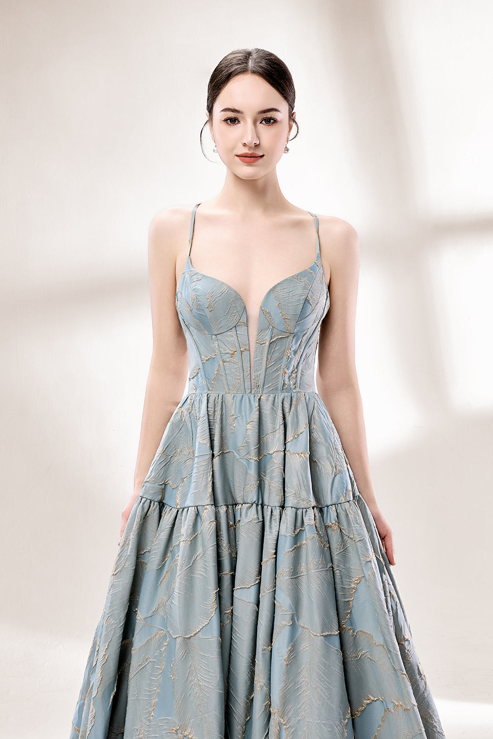 A woman stands against a light background, wearing a light - blue, tiered gown with gold - toned feather patterns. It has thin straps, a deep V - neckline, and a voluminous skirt. The unique jacquard design and elegant silhouette exude romance, perfect for formal events like garden parties or daytime weddings.5