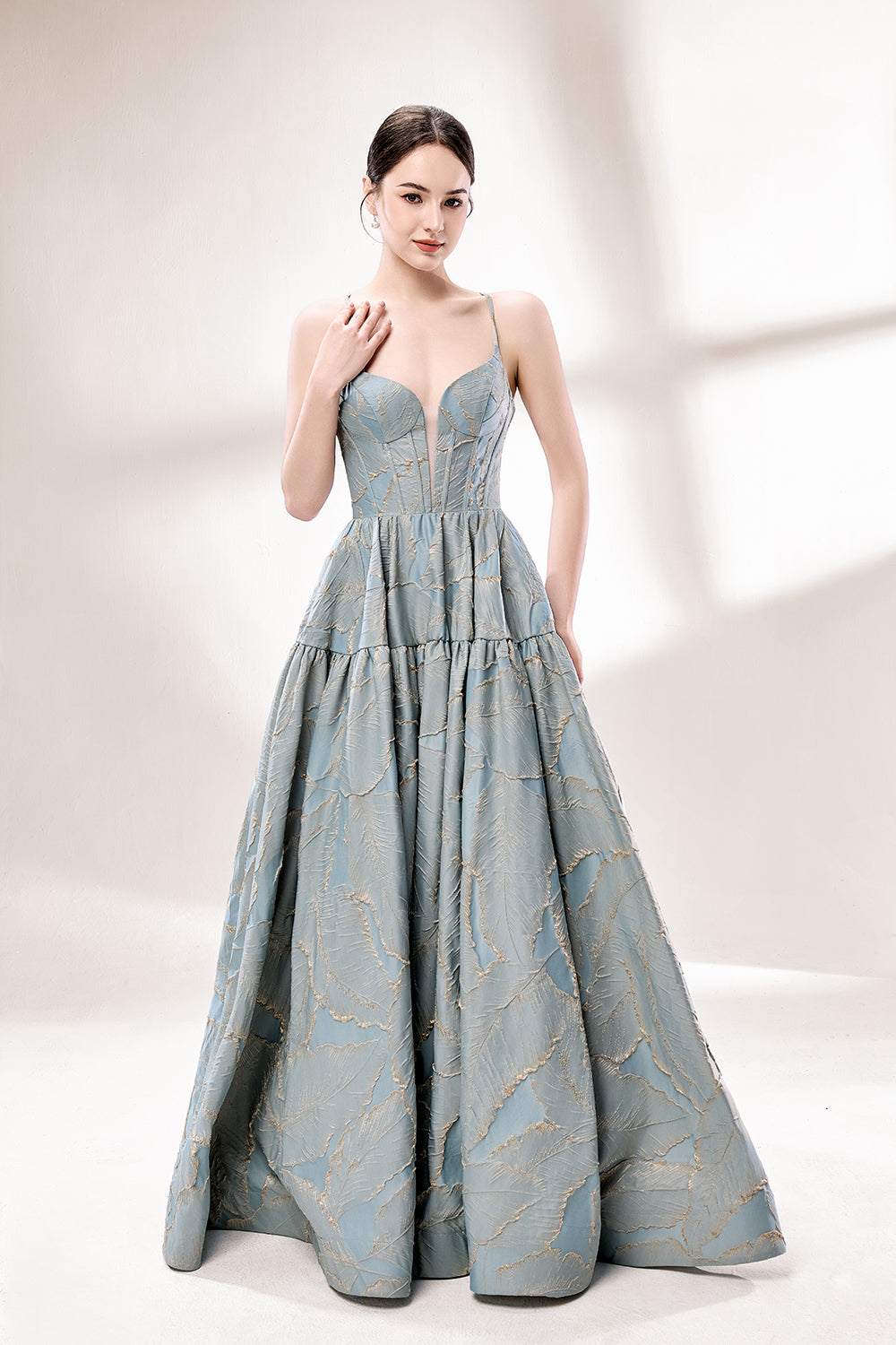 A woman stands against a light background, wearing a light - blue, tiered gown with gold - toned feather patterns. It has thin straps, a deep V - neckline, and a voluminous skirt. The unique jacquard design and elegant silhouette exude romance, perfect for formal events like garden parties or daytime weddings.4