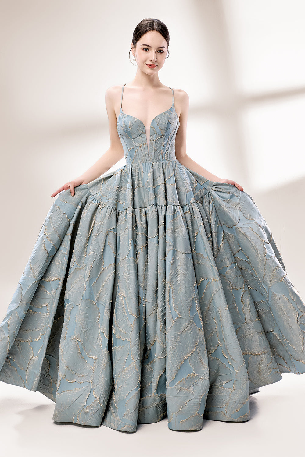 A woman stands against a light background, wearing a light - blue, tiered gown with gold - toned feather patterns. It has thin straps, a deep V - neckline, and a voluminous skirt. The unique jacquard design and elegant silhouette exude romance, perfect for formal events like garden parties or daytime weddings.1