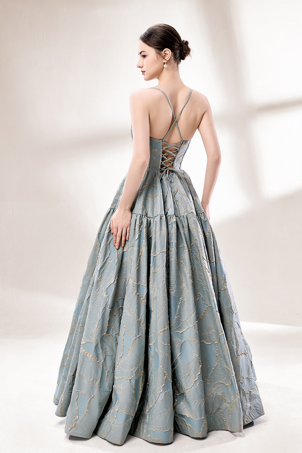 A woman stands against a light background, wearing a light - blue, tiered gown with gold - toned feather patterns. It has thin straps, a deep V - neckline, and a voluminous skirt. The unique jacquard design and elegant silhouette exude romance, perfect for formal events like garden parties or daytime weddings.2