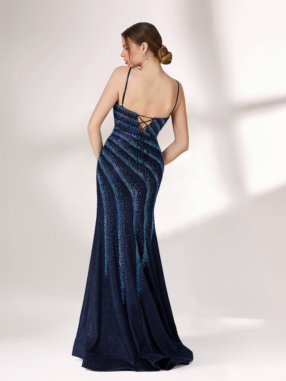 A woman stands against a light background, wearing a navy blue evening gown. It features thin spaghetti straps, a deep V - neckline, and diagonal stripes of shimmering blue embellishments. The dress has a high side slit, creating an elegant and sexy look, perfect for formal events.4