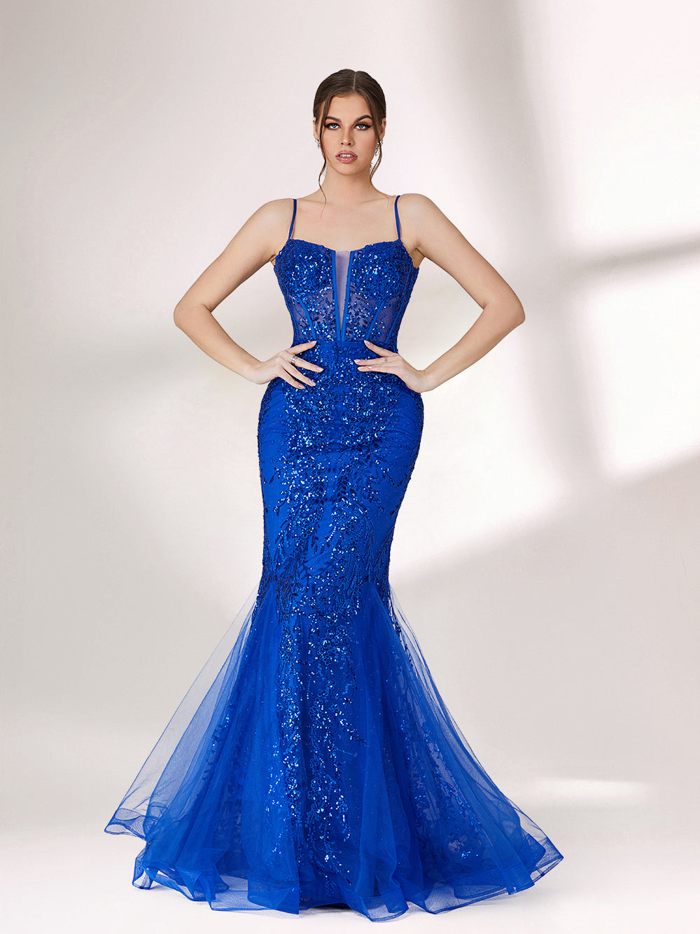 A woman stands against a light background, wearing a bright blue mermaid - style evening gown. It features thin spaghetti straps, a sheer panel at the neckline, and is covered in sparkling sequins. The tulle skirt flares out elegantly, creating a glamorous look perfect for formal events.3