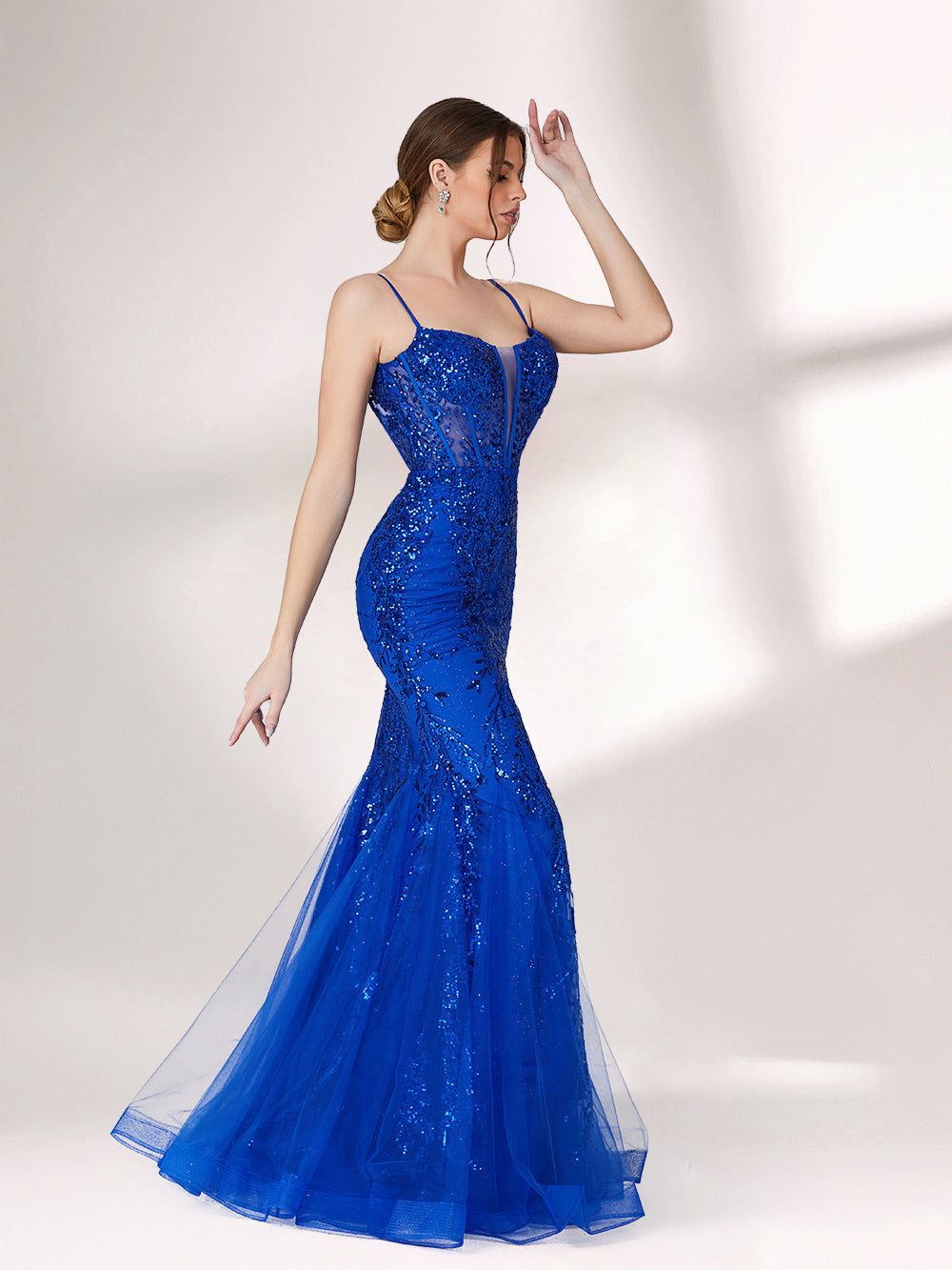 A woman stands against a light background, wearing a bright blue mermaid - style evening gown. It features thin spaghetti straps, a sheer panel at the neckline, and is covered in sparkling sequins. The tulle skirt flares out elegantly, creating a glamorous look perfect for formal events.5