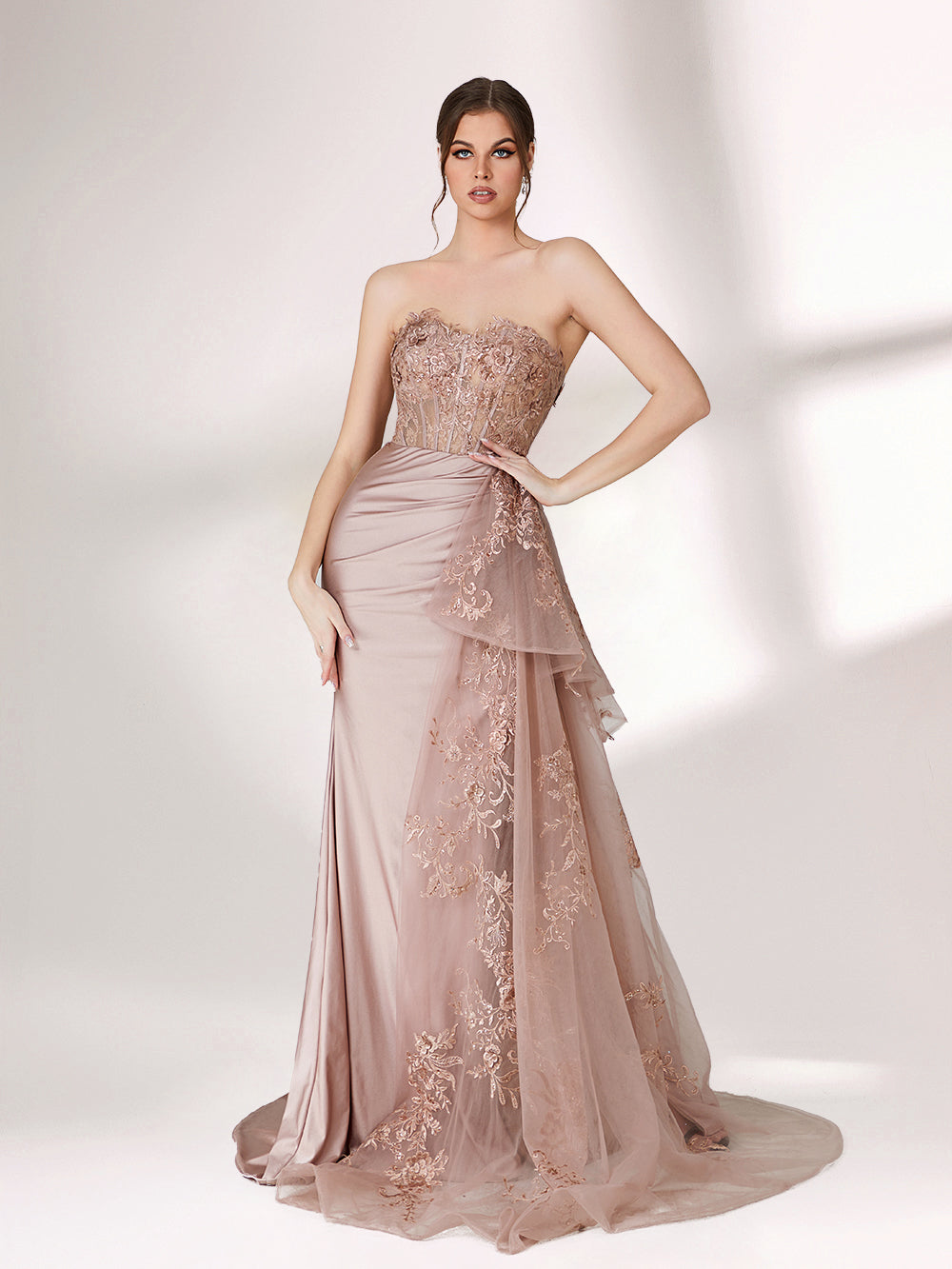 A woman stands against a light background, wearing a blush - toned strapless evening gown. The bodice features intricate floral embroidery, and there's a draped, embroidered tulle overlay on the skirt with a small train. The design is elegant and romantic, suitable for formal events.1