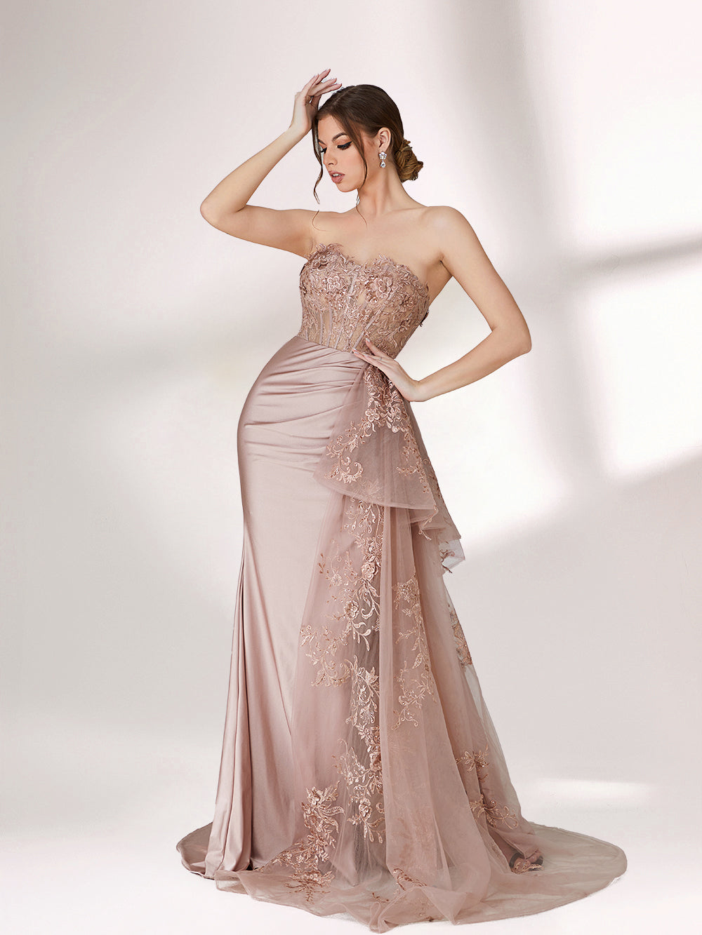 A woman stands against a light background, wearing a blush - toned strapless evening gown. The bodice features intricate floral embroidery, and there's a draped, embroidered tulle overlay on the skirt with a small train. The design is elegant and romantic, suitable for formal events.3