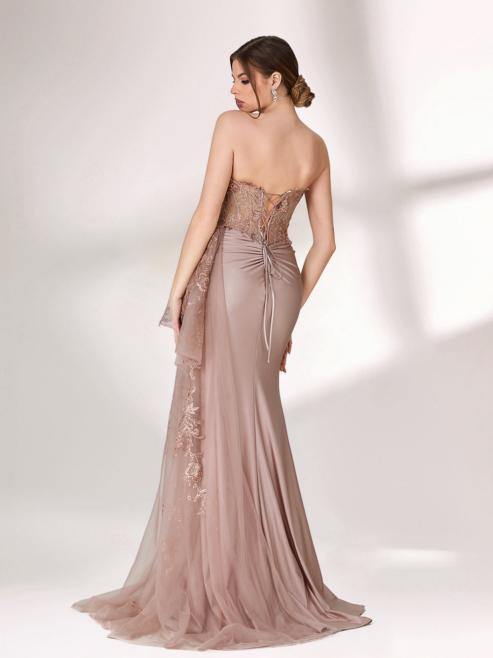 A woman stands against a light background, wearing a blush - toned strapless evening gown. The bodice features intricate floral embroidery, and there's a draped, embroidered tulle overlay on the skirt with a small train. The design is elegant and romantic, suitable for formal events.2