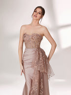 A woman stands against a light background, wearing a blush - toned strapless evening gown. The bodice features intricate floral embroidery, and there's a draped, embroidered tulle overlay on the skirt with a small train. The design is elegant and romantic, suitable for formal events.5