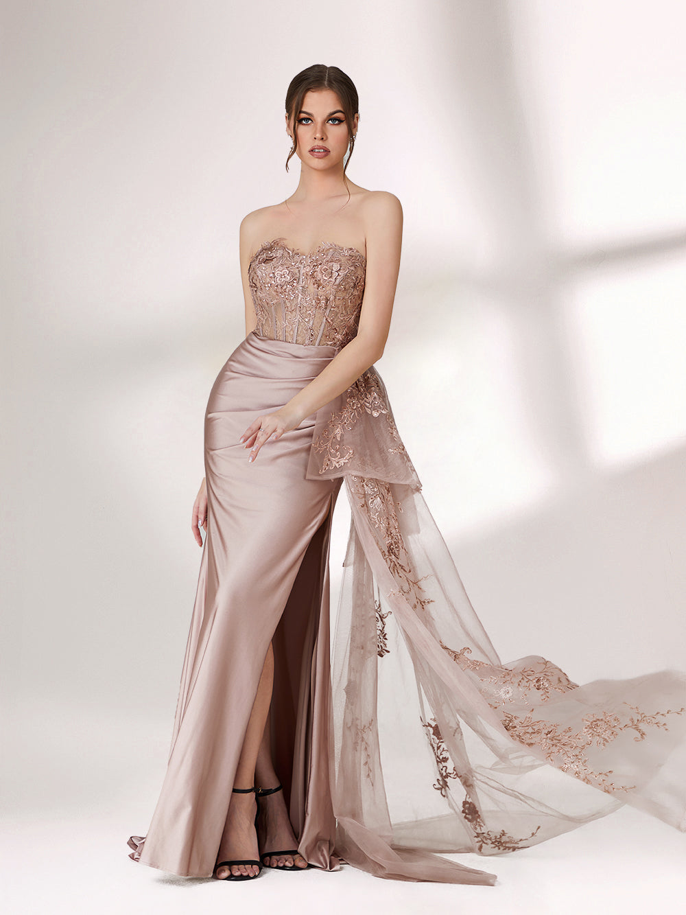 A woman stands against a light background, wearing a blush - toned strapless evening gown. The bodice features intricate floral embroidery, and there's a draped, embroidered tulle overlay on the skirt with a small train. The design is elegant and romantic, suitable for formal events.7